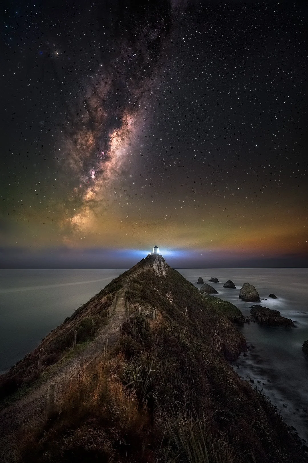 A lighthouse on a hill at night with the Milky Way visible in the sky above.