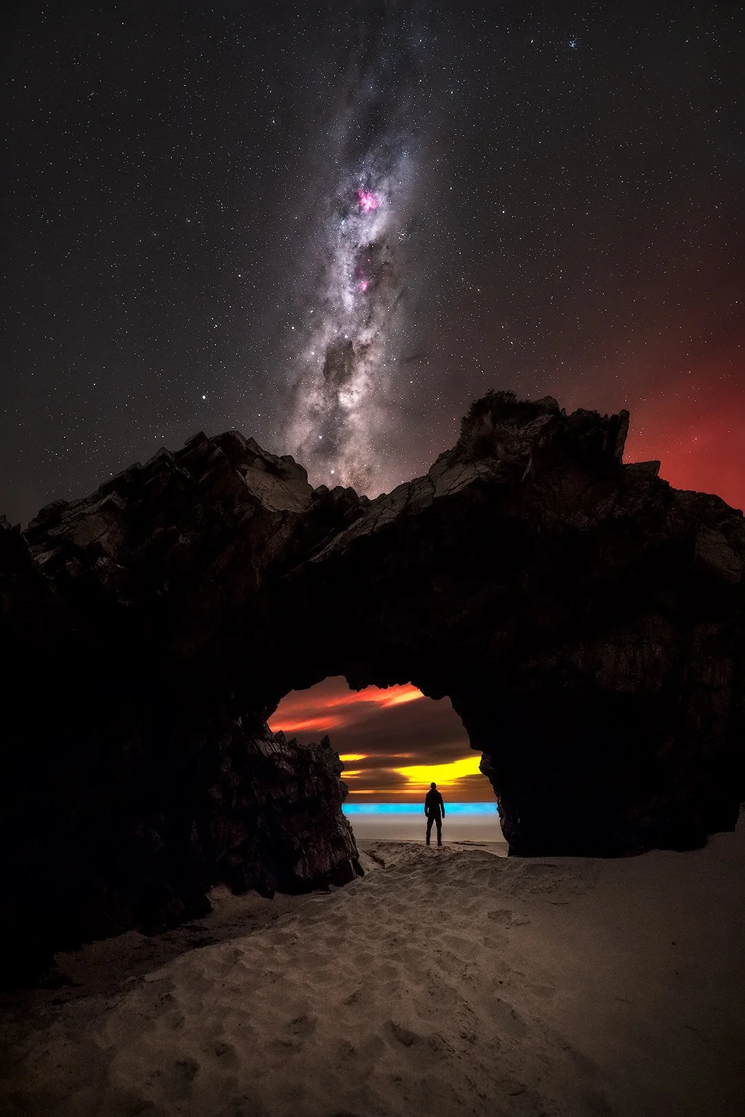 A person standing on a sandy beach, silhouetted against a colorful sunset sky seen through a natural rock formation arch, with a starry night sky and the Milky Way galaxy overhead.