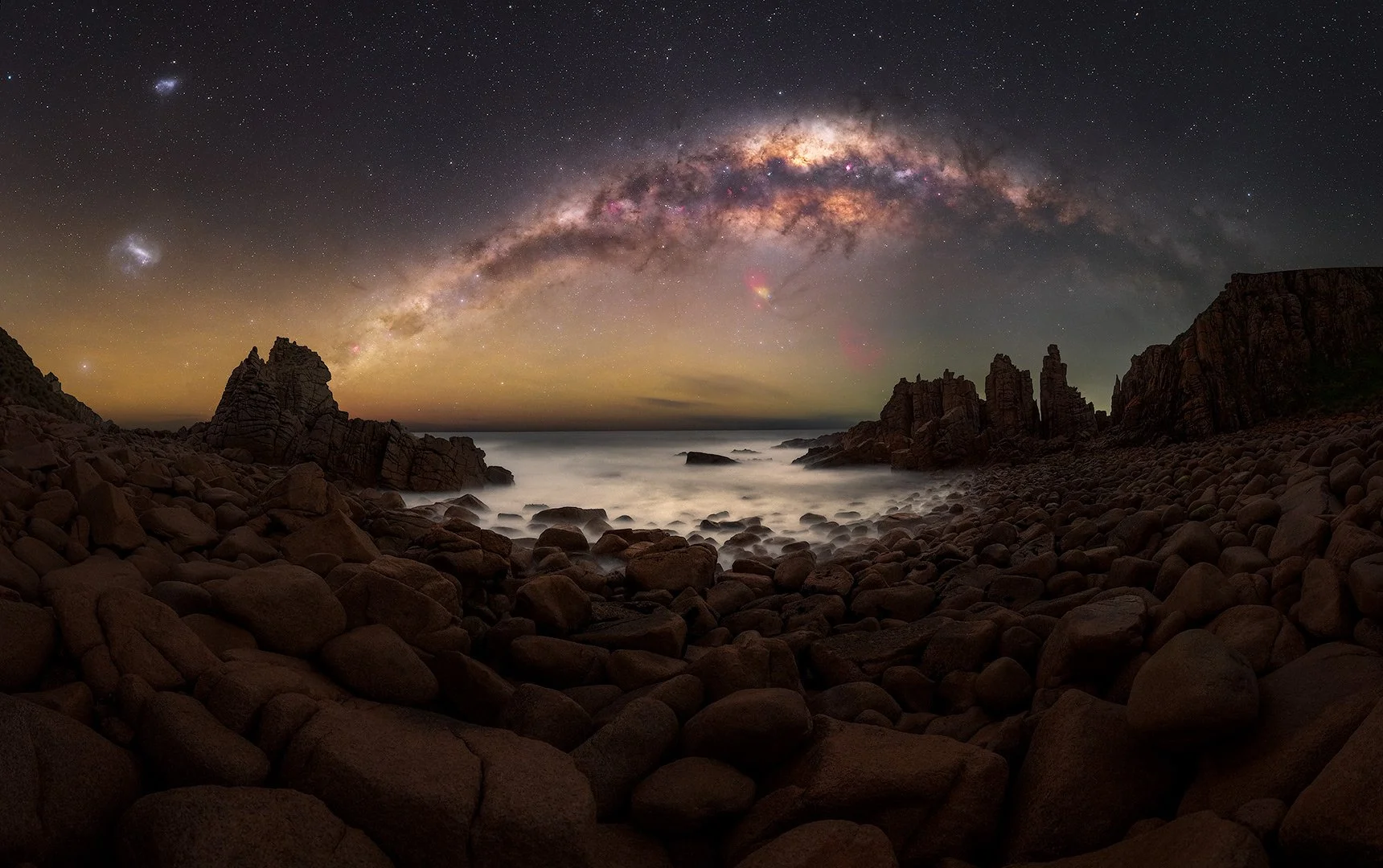 Nighttime coastal landscape with rocky shore under a star-filled sky and the Milky Way galaxy arched overhead.