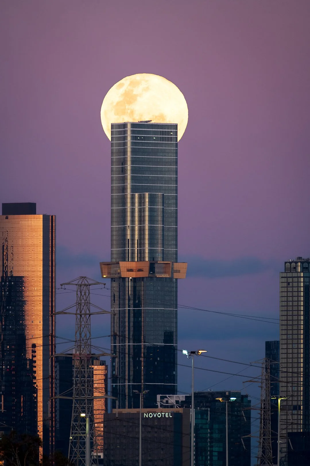 City skyline at dusk with tall buildings and a large full moon behind one skyscraper.