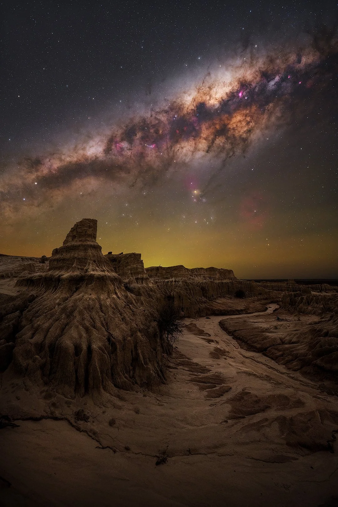 Nighttime landscape with rugged desert terrain and rock formations in the foreground under a starry sky. The Milky Way galaxy is prominently visible overhead with bright stars and colorful nebulae.