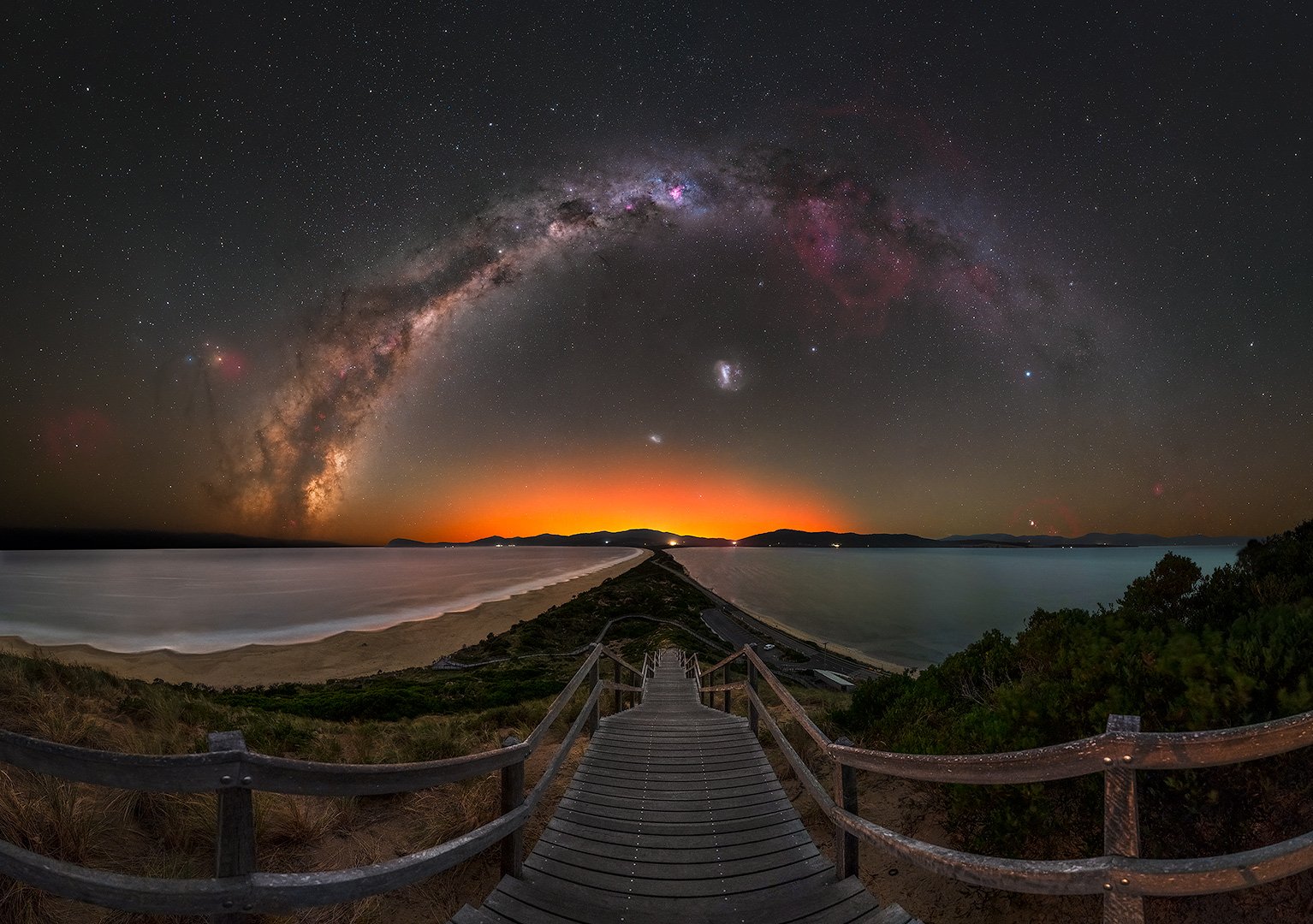 A wooden walkway leading to the beach at sunset with the Milky Way galaxy visible in the night sky, surrounded by stars, clouds, and a distant horizon