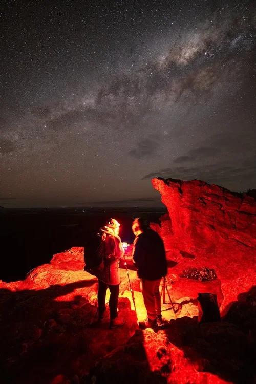 Two people with backpacks and hiking gear standing on a rocky terrain at night, illuminated by red light, under a starry sky with the Milky Way visible, near a red rock formation.