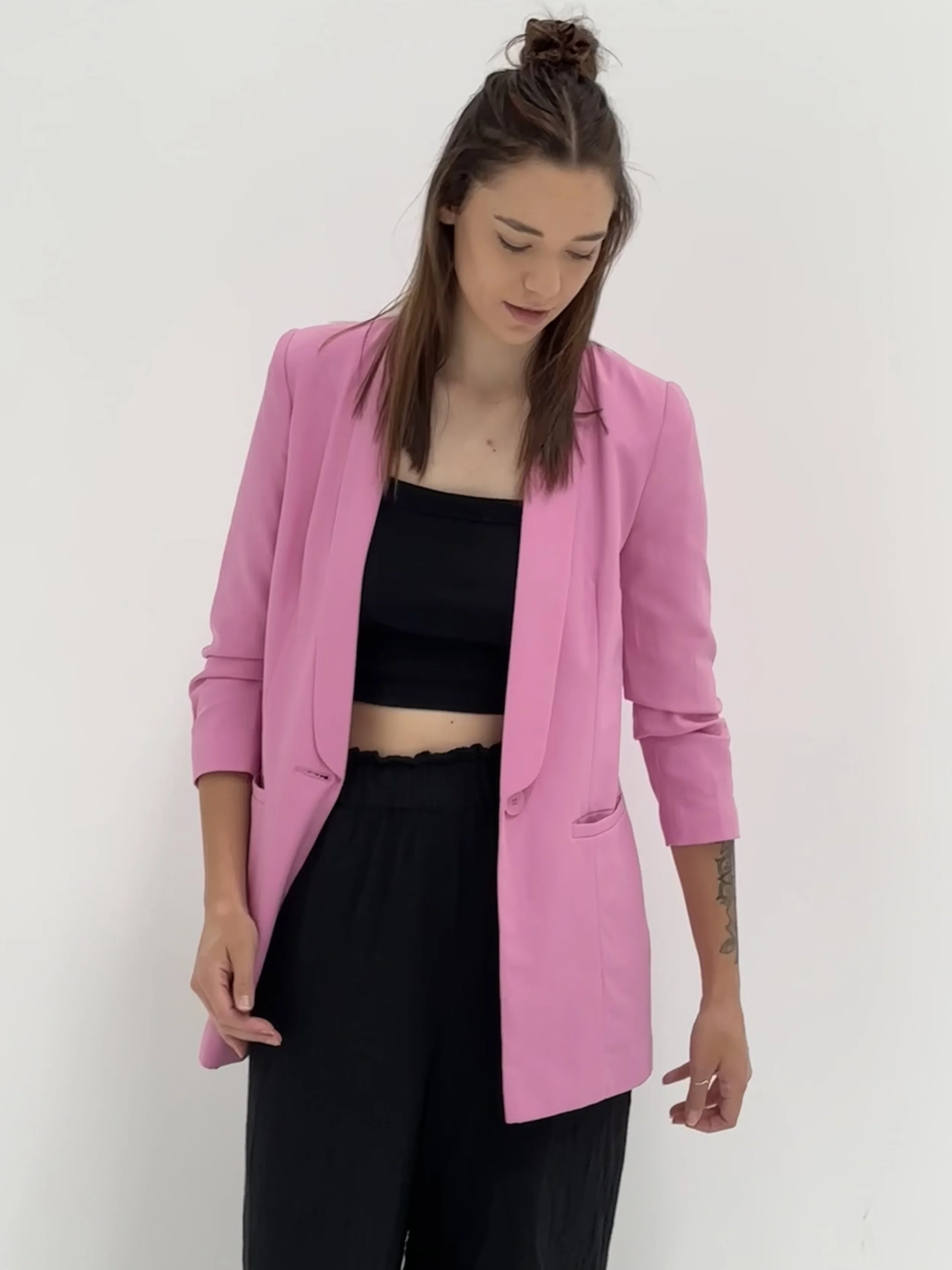 Sarah, the founder, wearing a pink blazer over a black crop top and black pants, standing against a plain white background.