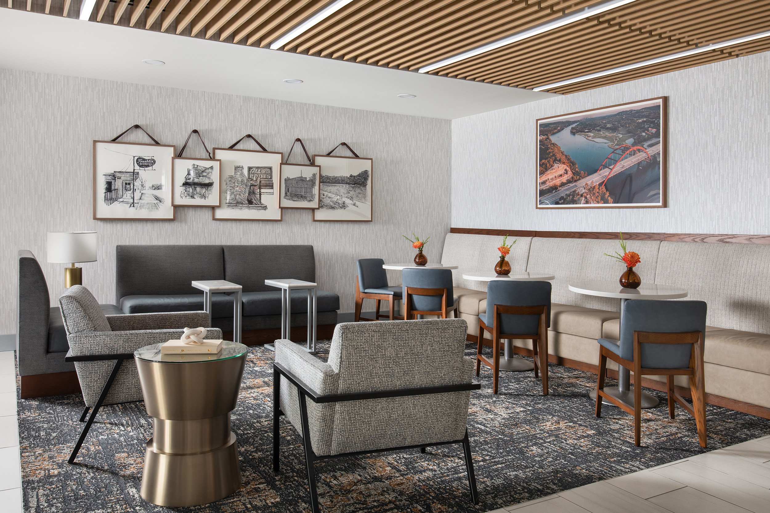 Modern hotel lobby with comfortable seating including armchairs, sofas, and small tables. Wall art features framed black-and-white sketches and a large photo of a bridge over a river. Decor includes vases with flowers and a patterned rug.