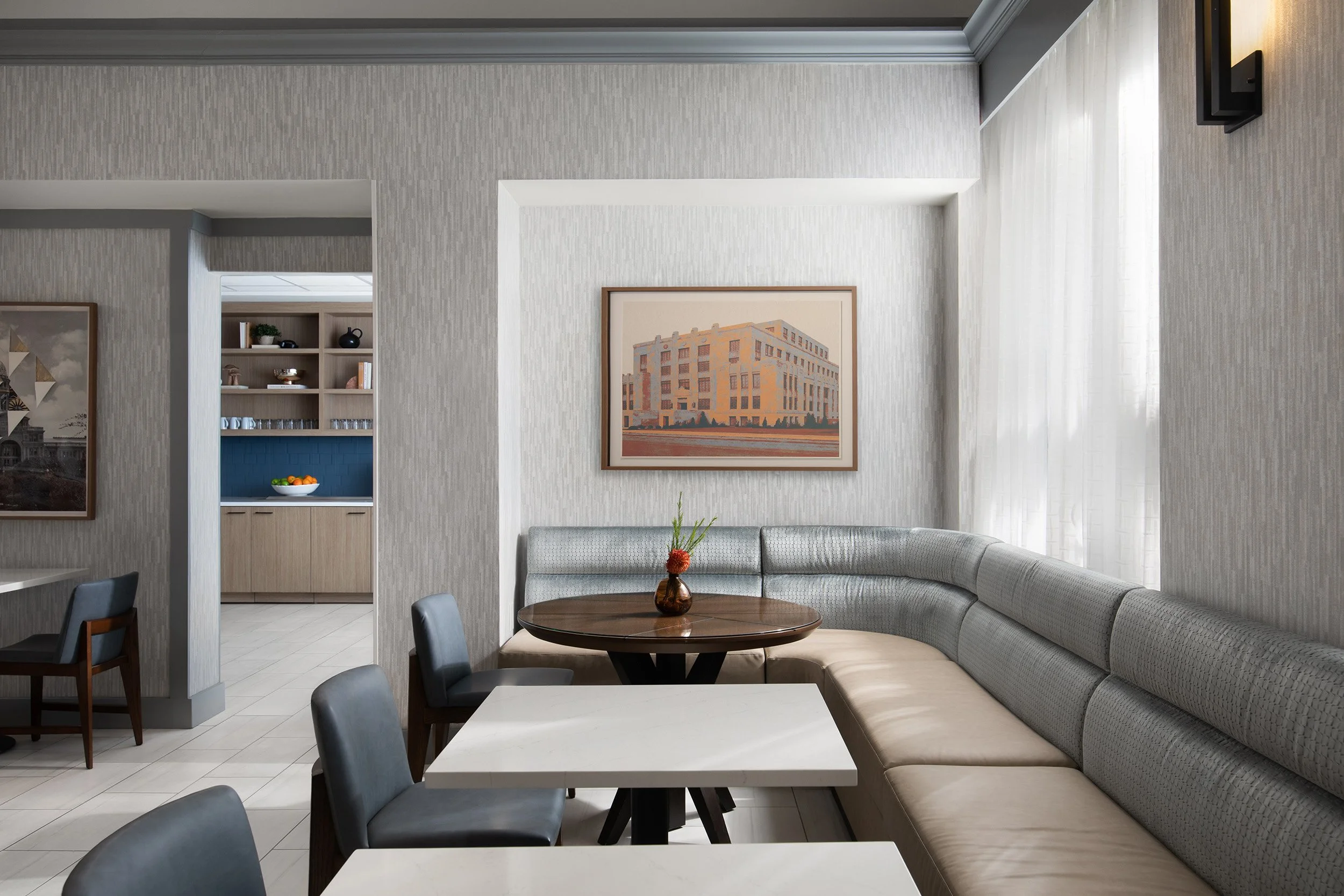 Interior of a modern dining area with a curved gray banquette, round wooden table with a small vase and flowers, white and gray chairs, and a framed building on the wall. In the background, a kitchen area with light wood cabinets and a bowl of oranges.