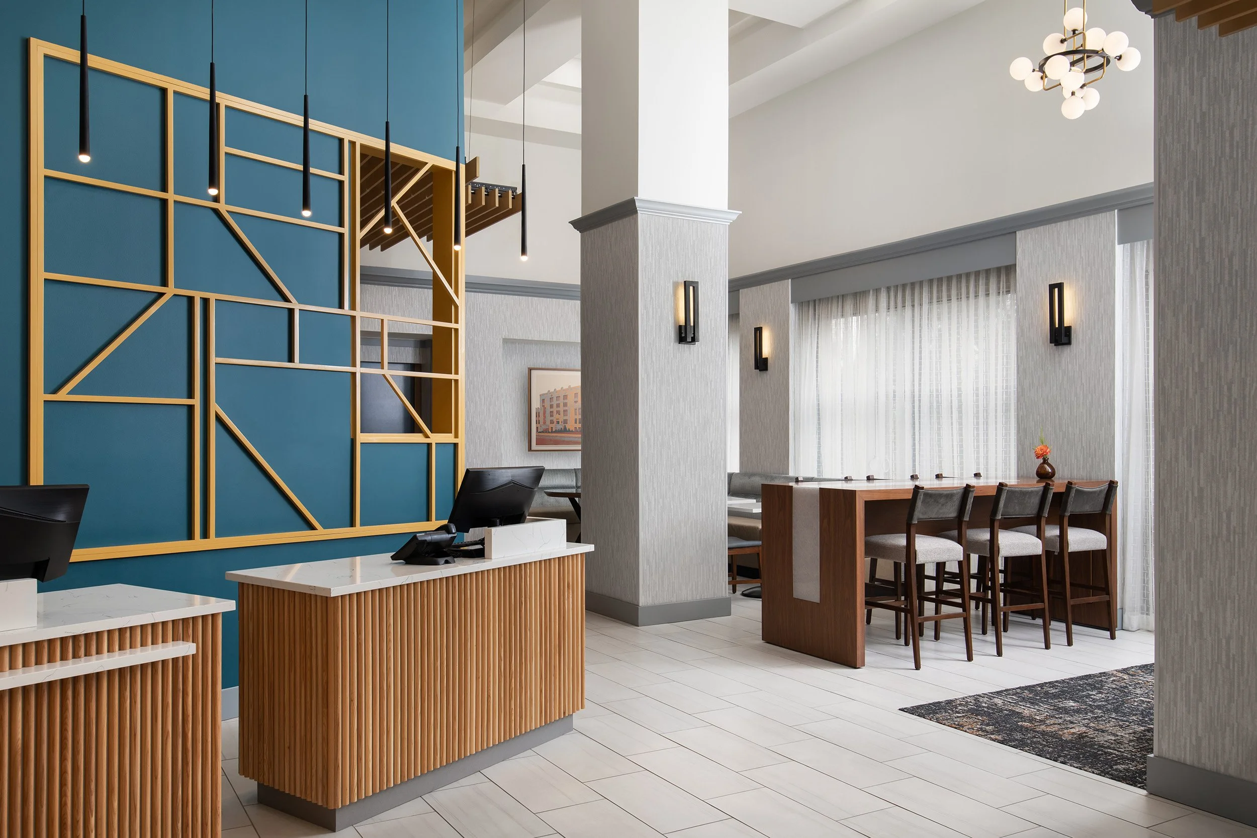 Modern hotel lobby with check-in counter, decorative blue wall with wood accents, reception desk with computers, seating area with high chairs, and large windows with sheer curtains.