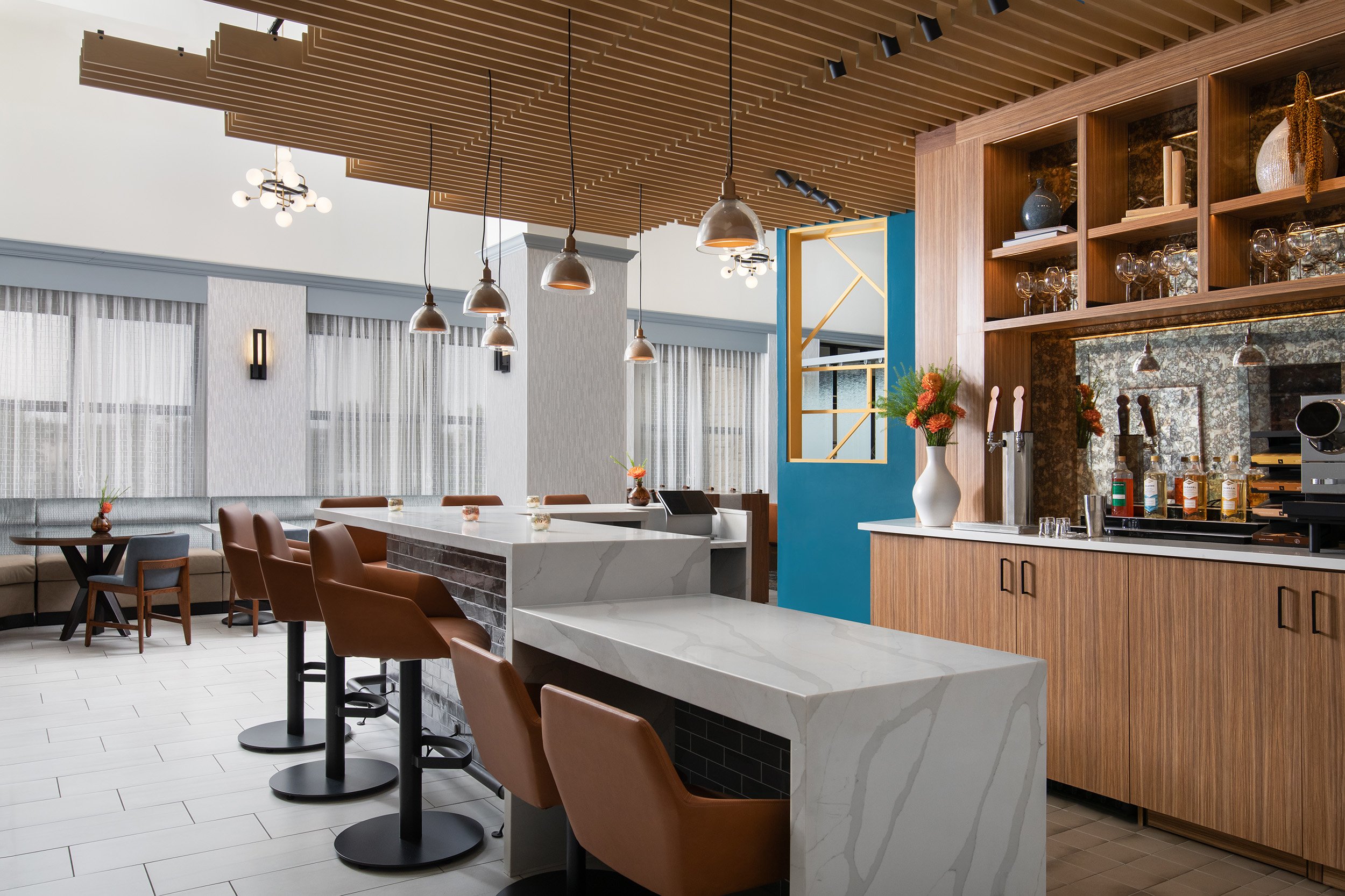 Interior view of a modern cafe or restaurant with a marble bar counter, brown leather bar stools, a wooden shelving unit with glassware, and decorative vases, featuring wood-paneled ceiling, pendant lighting, and large windows with sheer curtains.