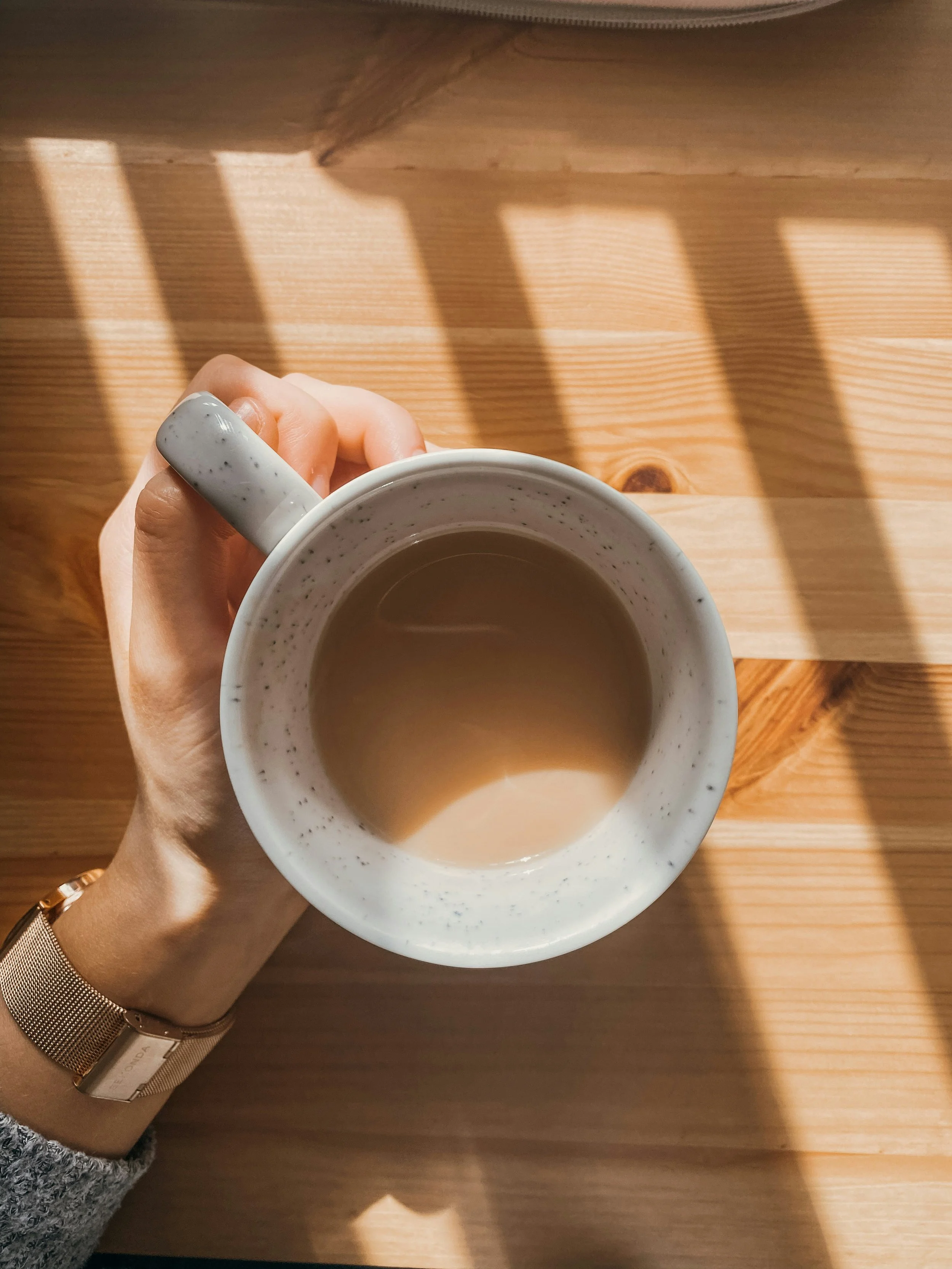 A person's hand holding a speckled white mug of coffee on a wooden table with striped sunlight shadows.