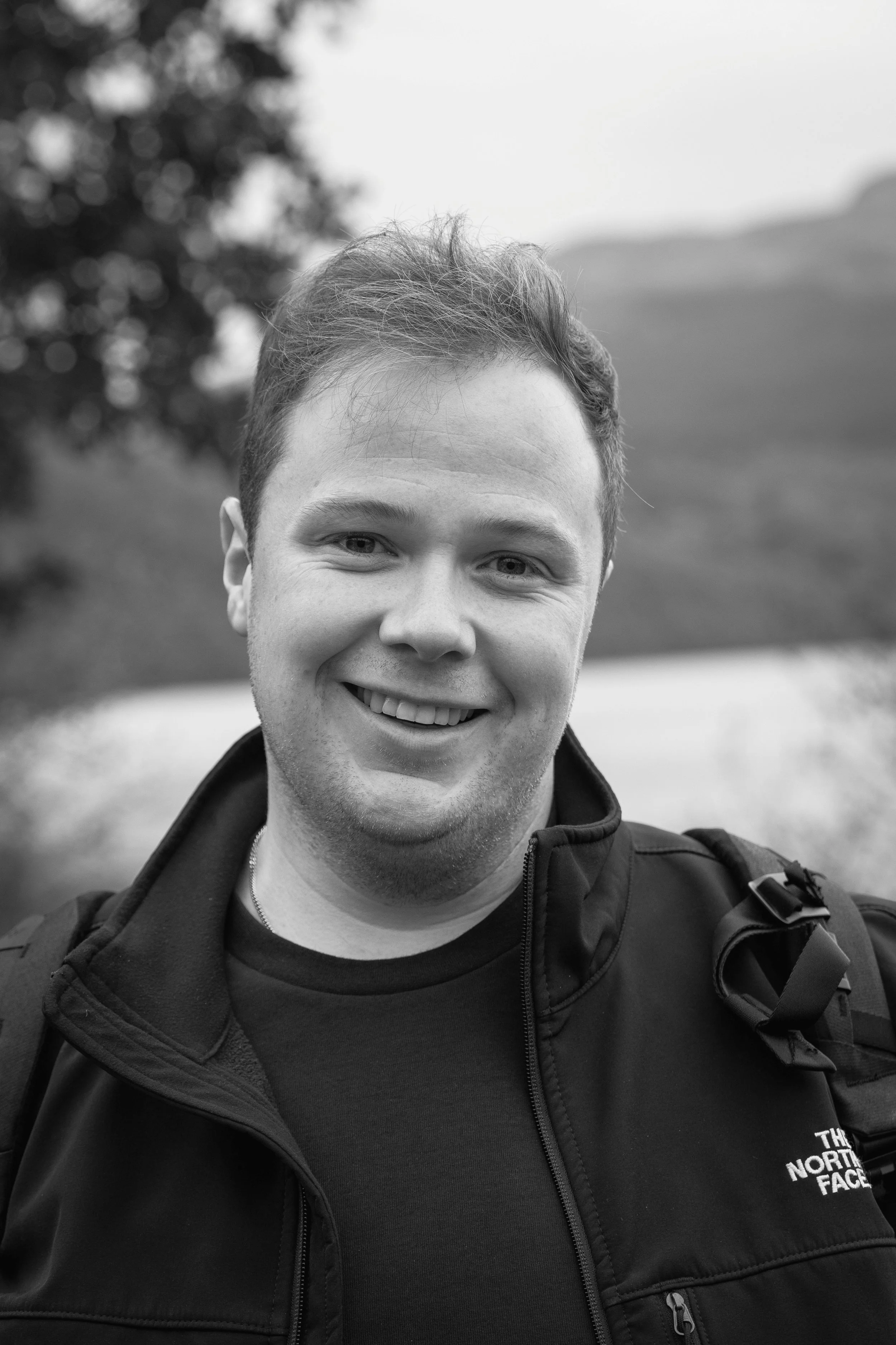 Smiling young man outdoors with a lake and trees in the background, wearing a black North Face jacket.