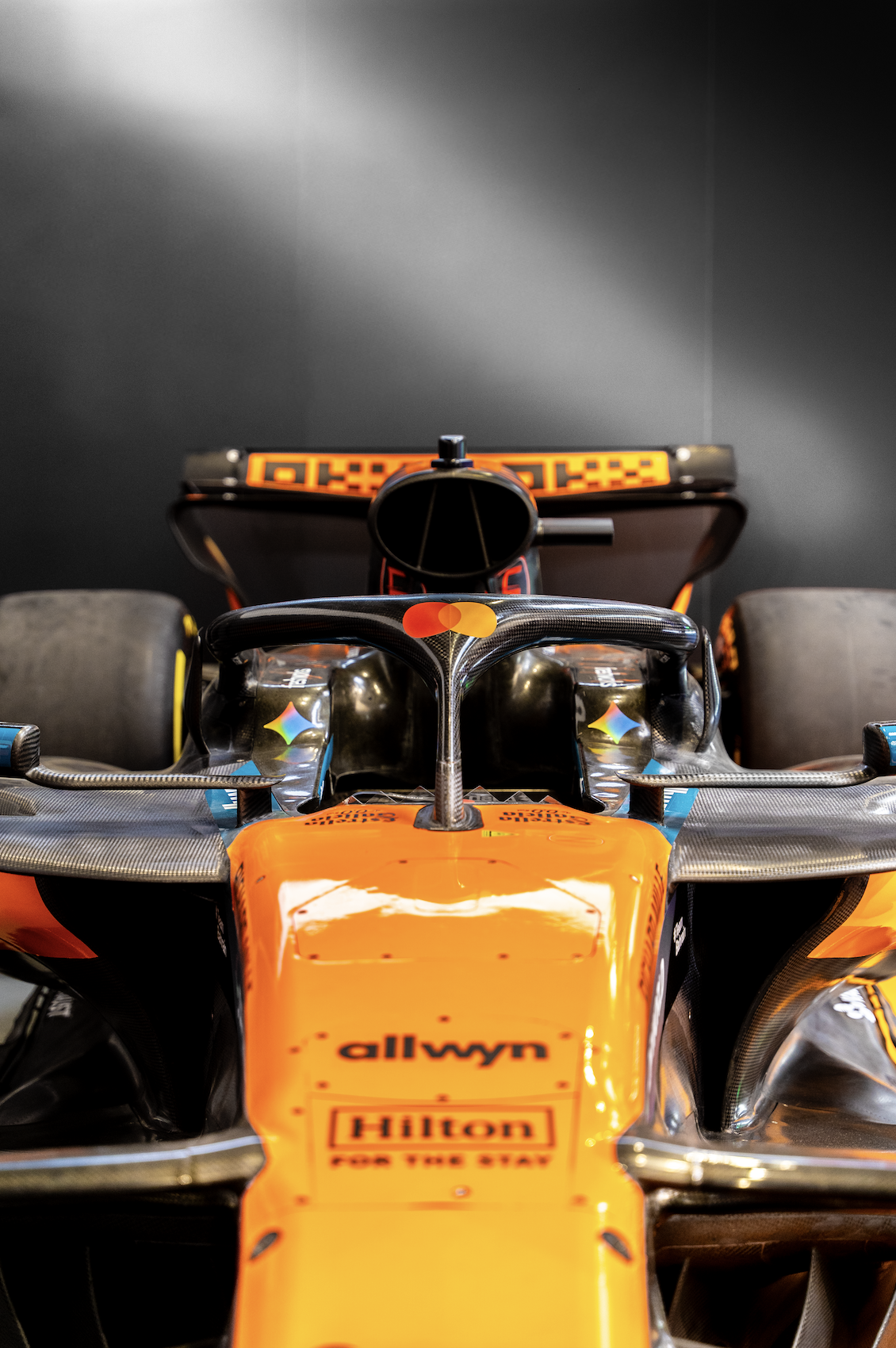 Front view of a Red Bull Racing Formula 1 car with orange accents, showcasing sponsor logos including Hack, Hilton, and allwyn, displayed in a dimly lit environment.