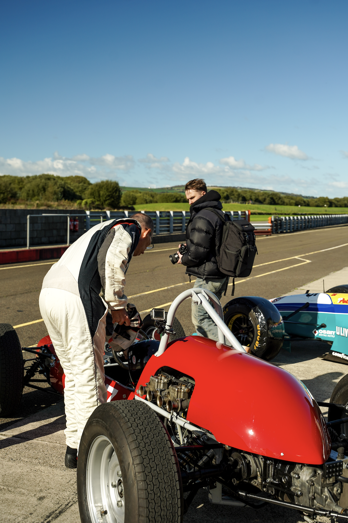 Two men in racing suits standing beside a red race car on a race track, one is working on the car while the other, holding a camera, looks on, with another race car parked nearby in a rural setting under a clear blue sky.