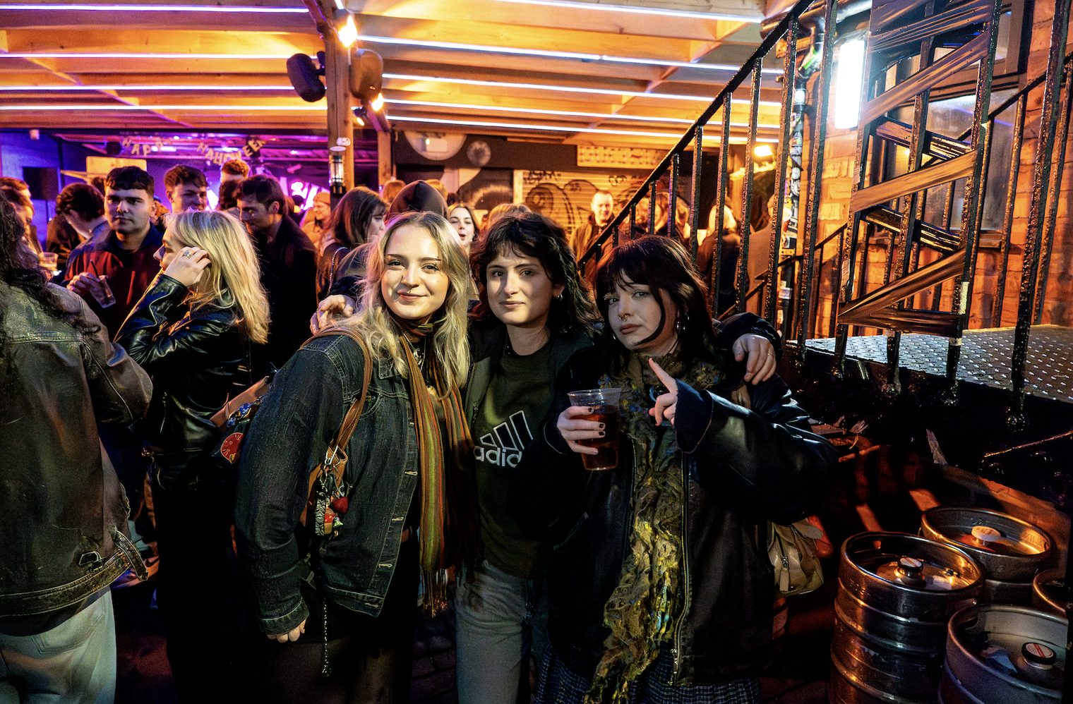 Group of people at a lively indoor event, three women in the foreground posing for a photo, one holding a drink, with colorful lighting and a bar area in the background.