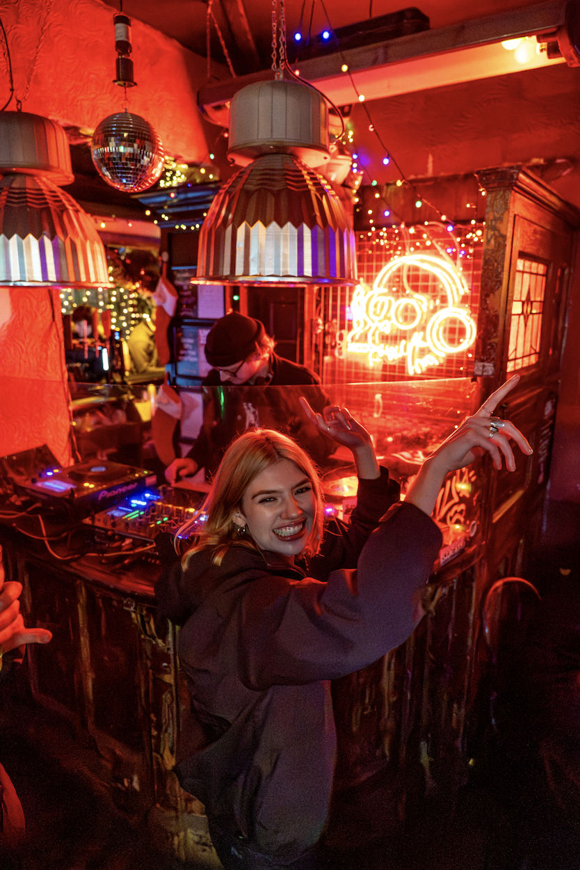 A woman smiling and pointing at the camera in a dimly lit, festive bar or club with neon lights, string lights, and a DJ in the background.