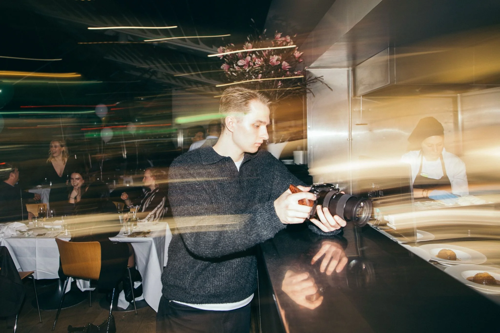 Young man with a camera in a busy restaurant or event space with blurred lights and guests in the background.