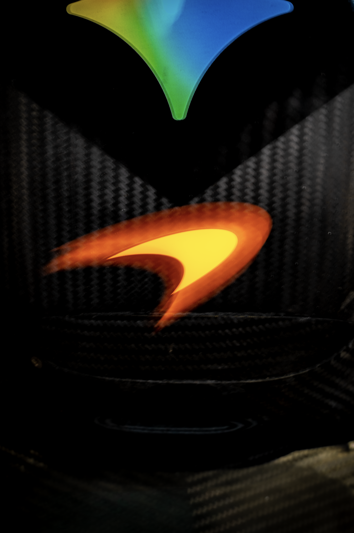 Close-up of a carbon fiber surface with illuminated colorful arrow-shaped logo in purple, blue, green, and orange on a black background.
