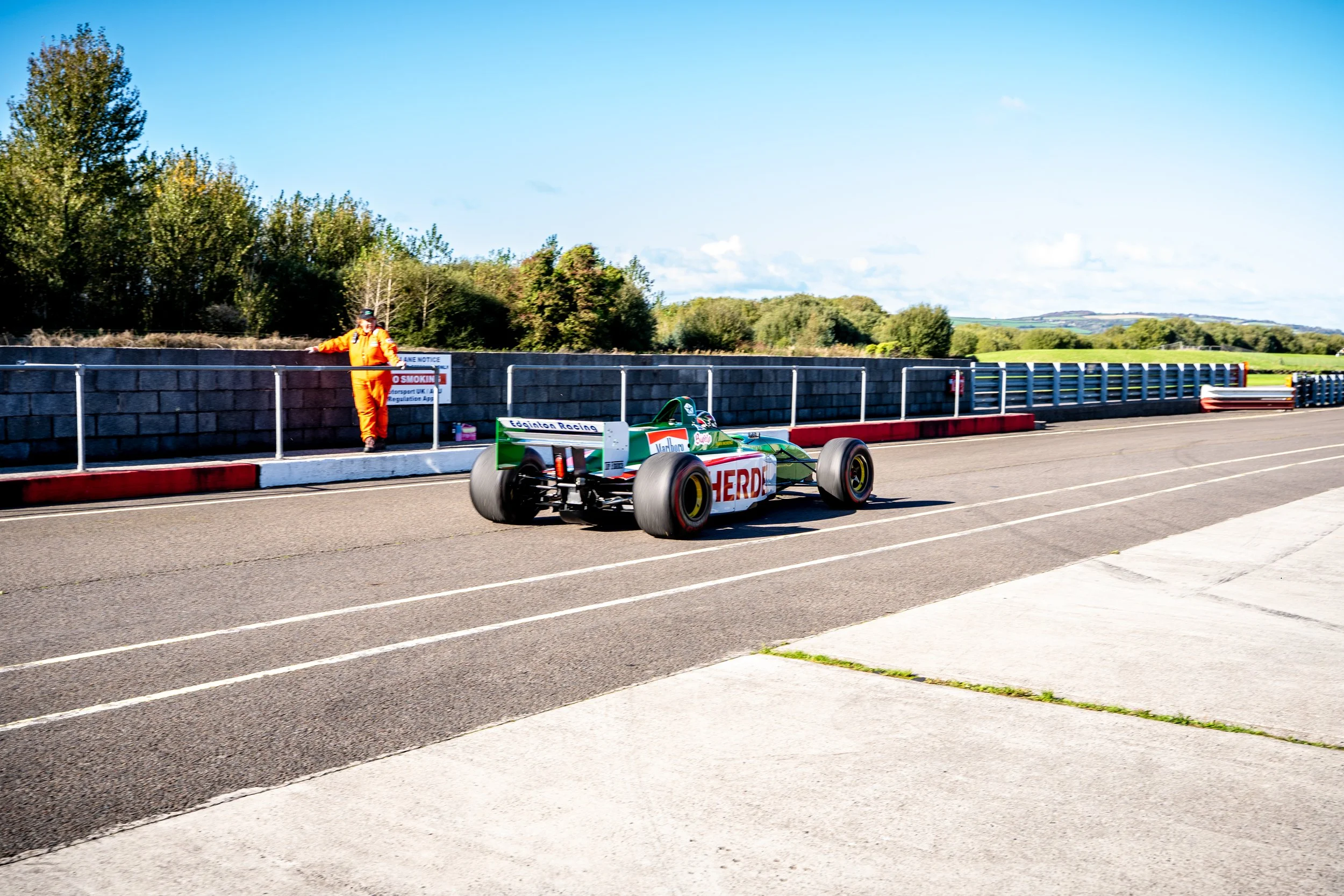A green and white race car on a track with a driver in a helmet, and a marshmallow in an orange jumpsuit standing behind a barrier on the side of the track with trees and blue sky in the background.
