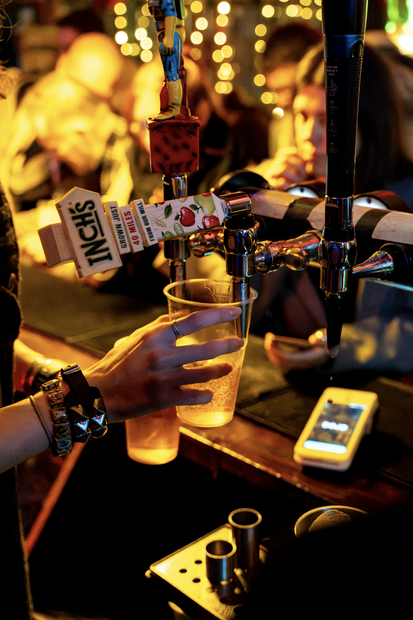 A person filling a clear plastic cup with beer from a tap at a bar, with several taps and people in the background during nighttime.
