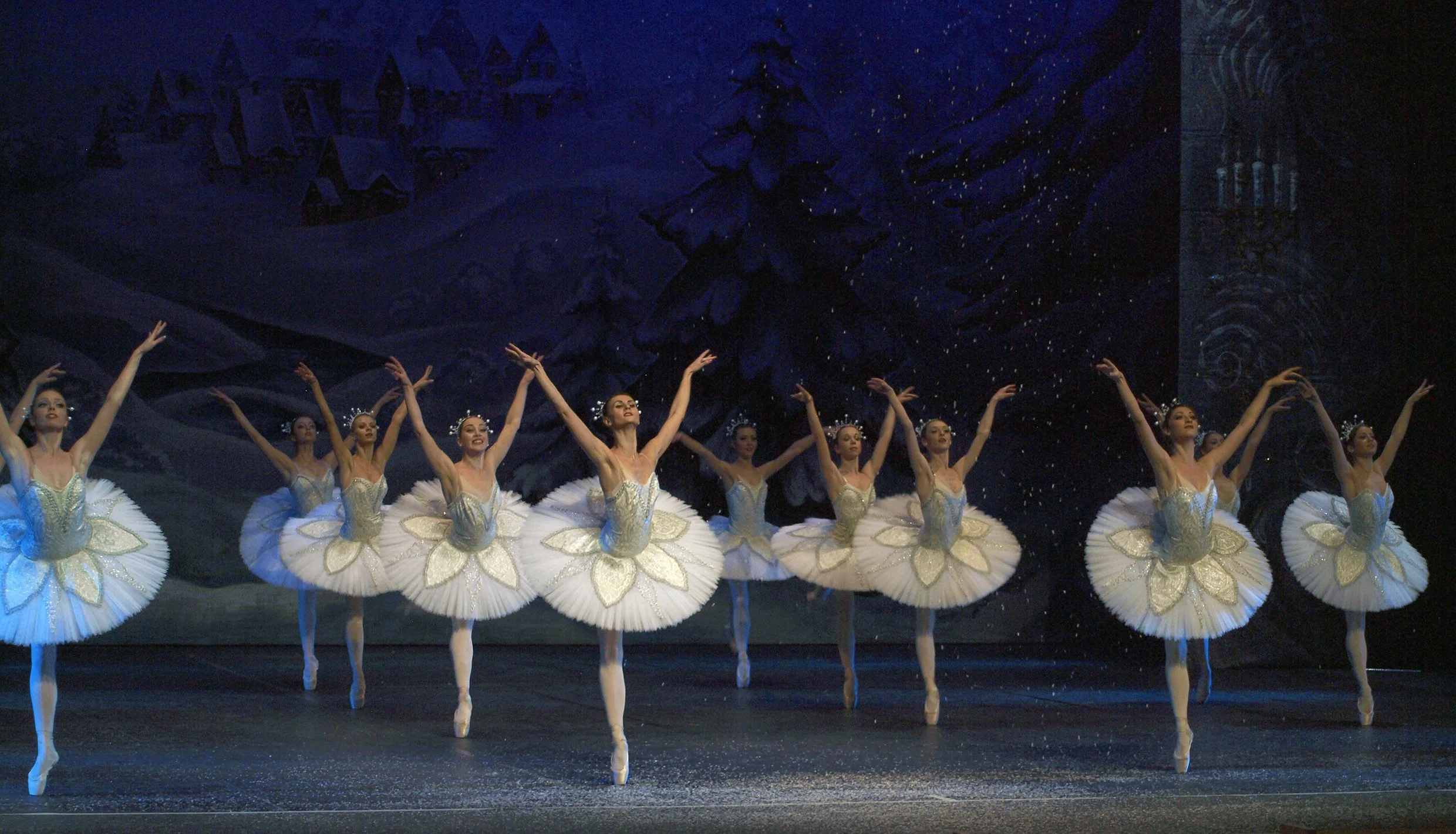 Grupo de bailarinas de ballet en un escenario, vestidas con tutús blancos decorados con copas de oro, realizando una danza clásica con fondo de decorado de invierno con árboles y un castillo.