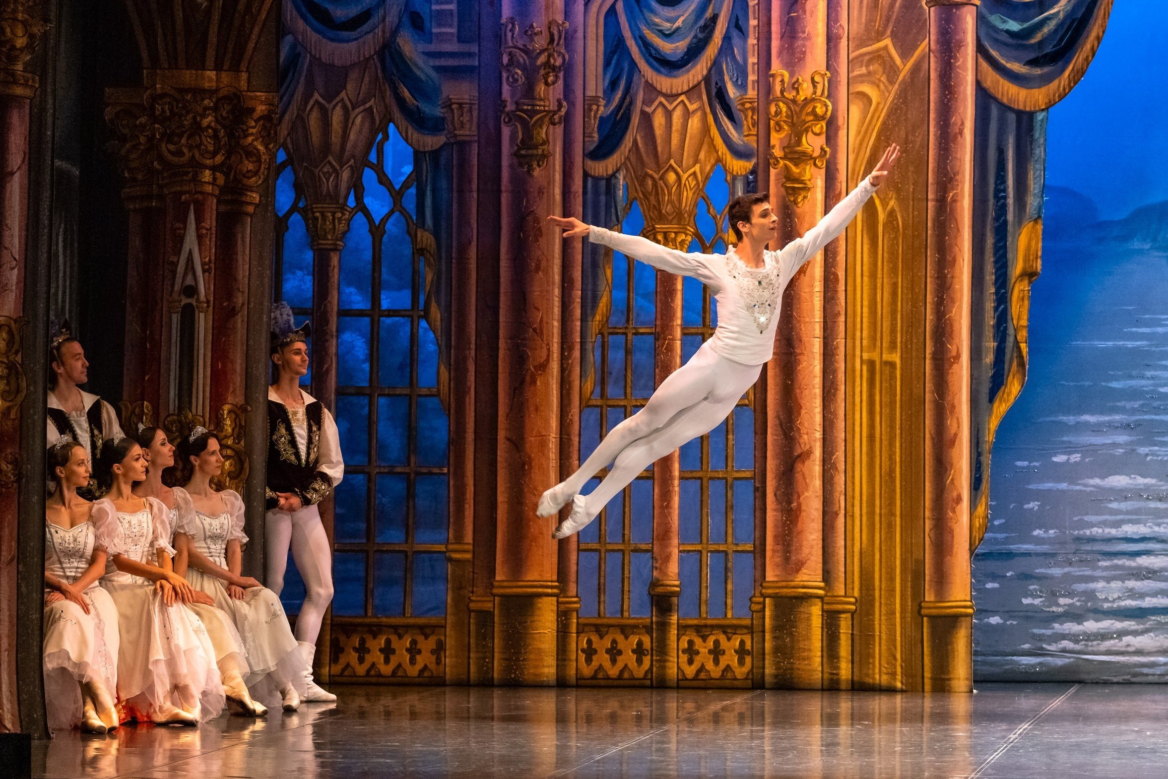 Bailarín masculino del Ballet Clásico Internacional en salto con un grupo de bailarinas y bailarines en el fondo en un escenario de estilo clásico con cortinas y ventanas decorativas.