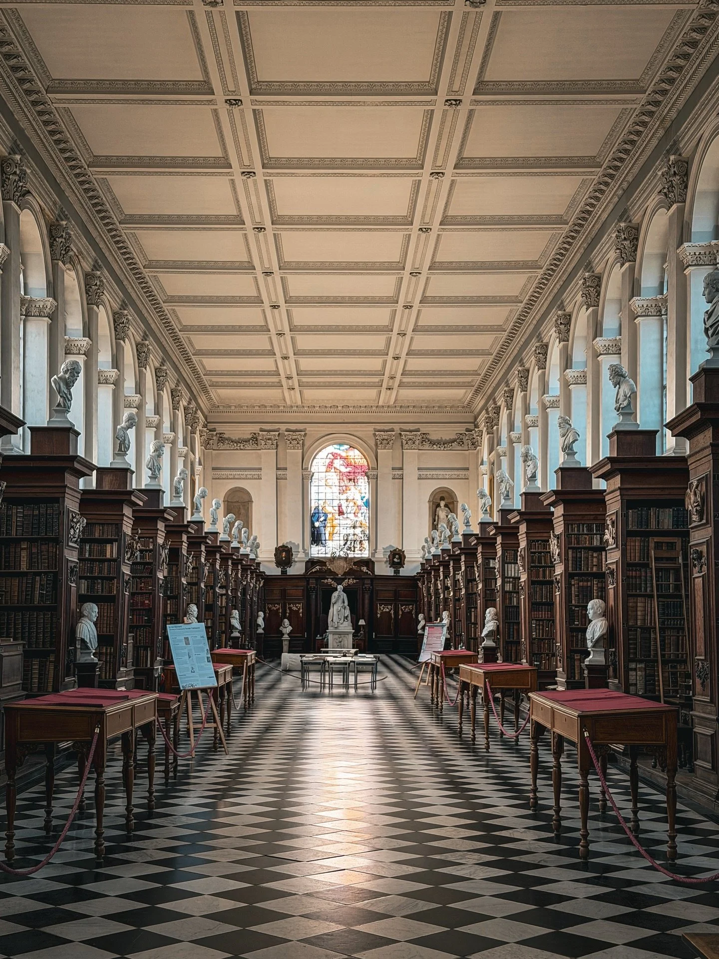 The interiors of the Wren Library at Trinity College are a masterclass in restrained classical elegance. Bathed in natural light from tall, evenly spaced windows, the long gallery feels both serene and purposeful. Dark timber bookcases line the walls
