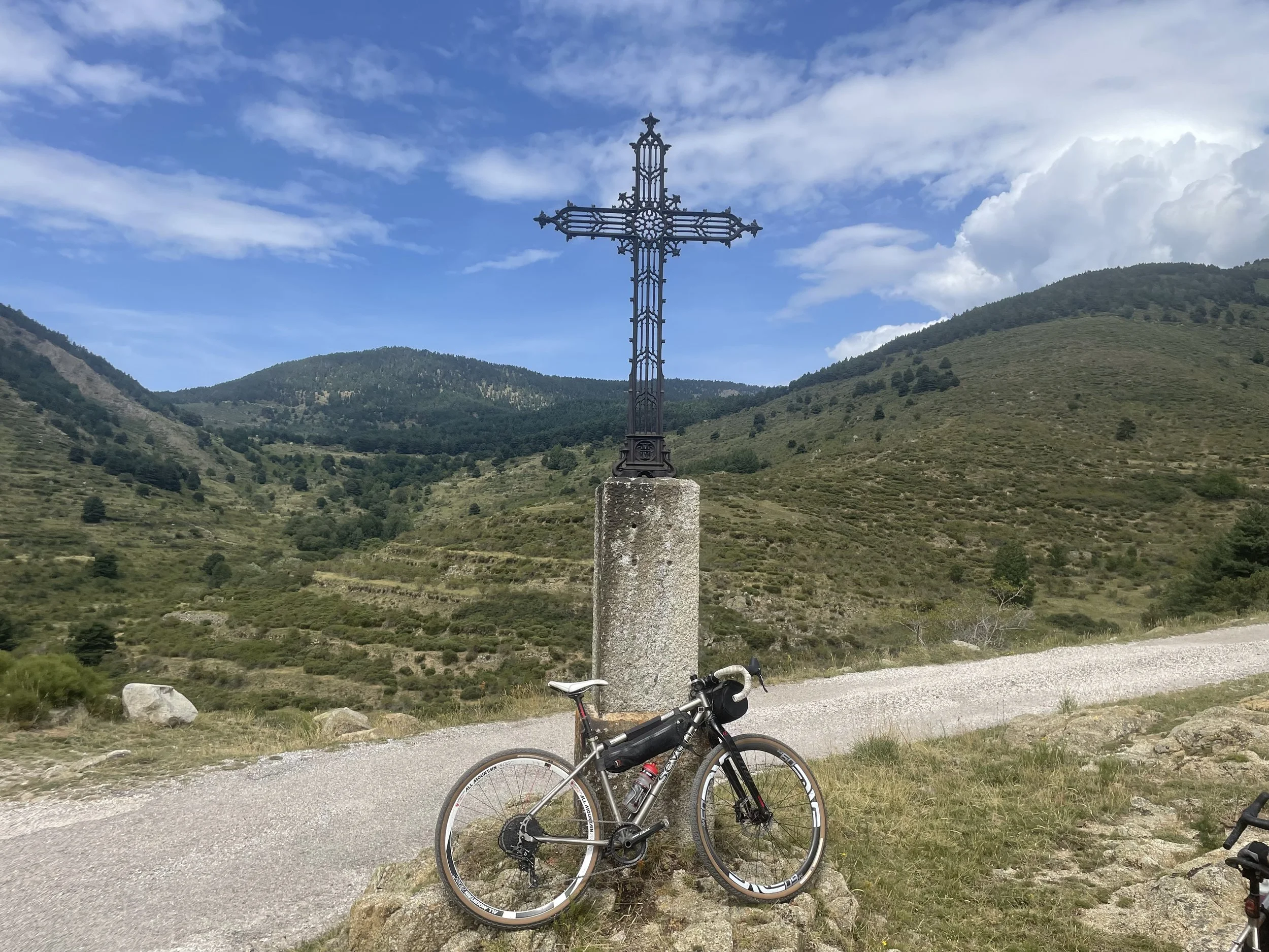 Gravel bike lent against an ironwork cross with mountains behind