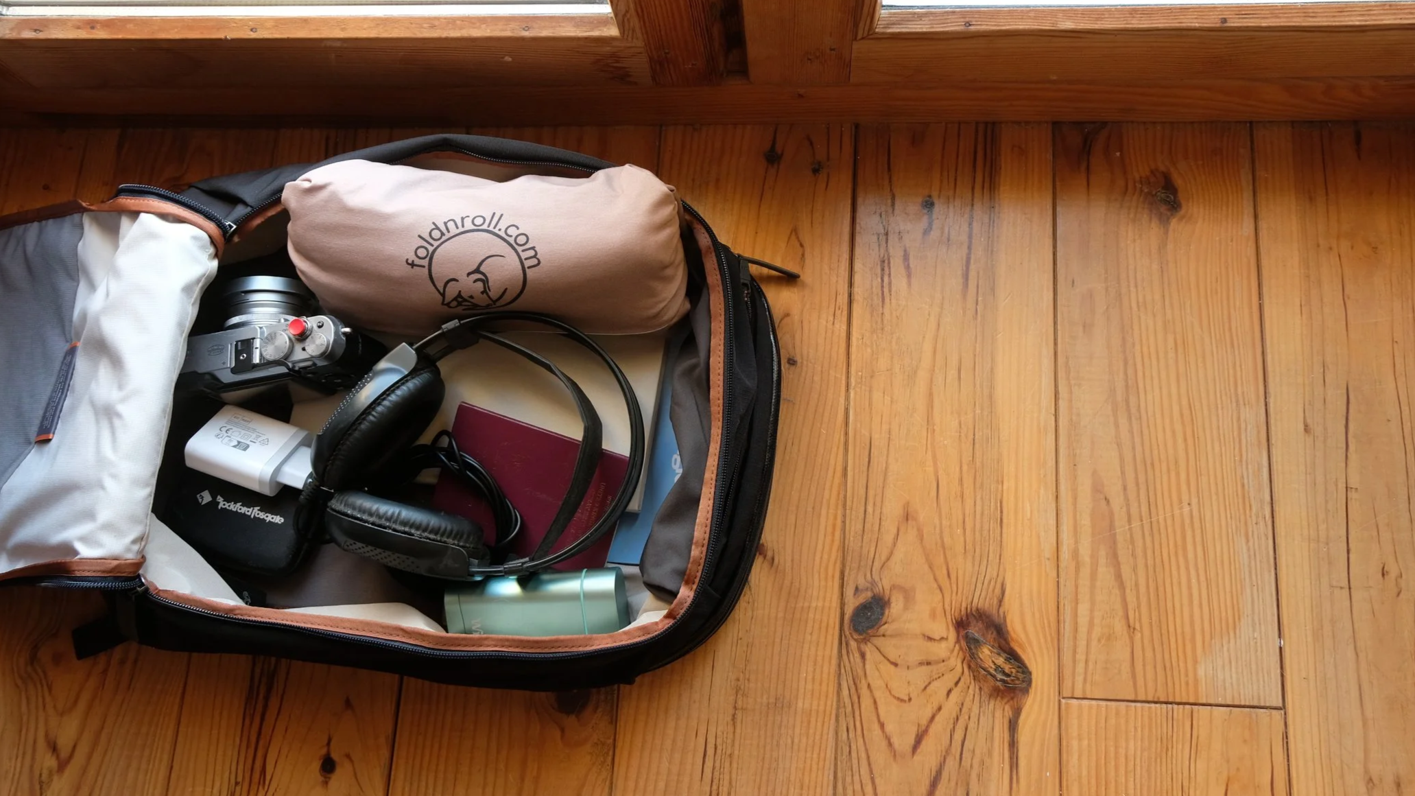 Open backpack with electronic accessories on wooden floor, including headphones, a portable speaker, a power bank, and a small beige bag with a logo.