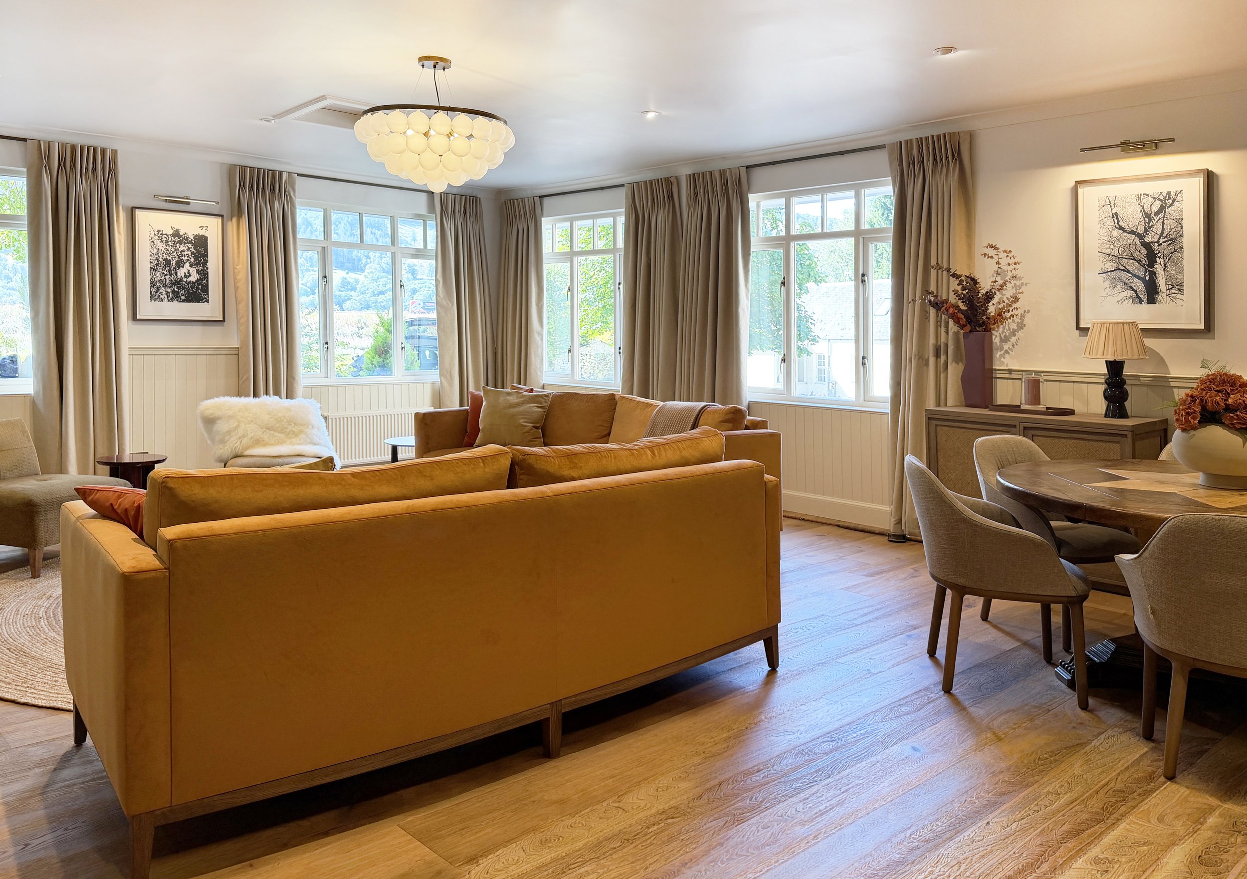 Living room with multiple large windows, beige curtains, a modern chandelier, a tan sofa with pillows, a cozy chair with a faux fur blanket, a side table, and artwork on the walls. East Haugh House Pitlochry