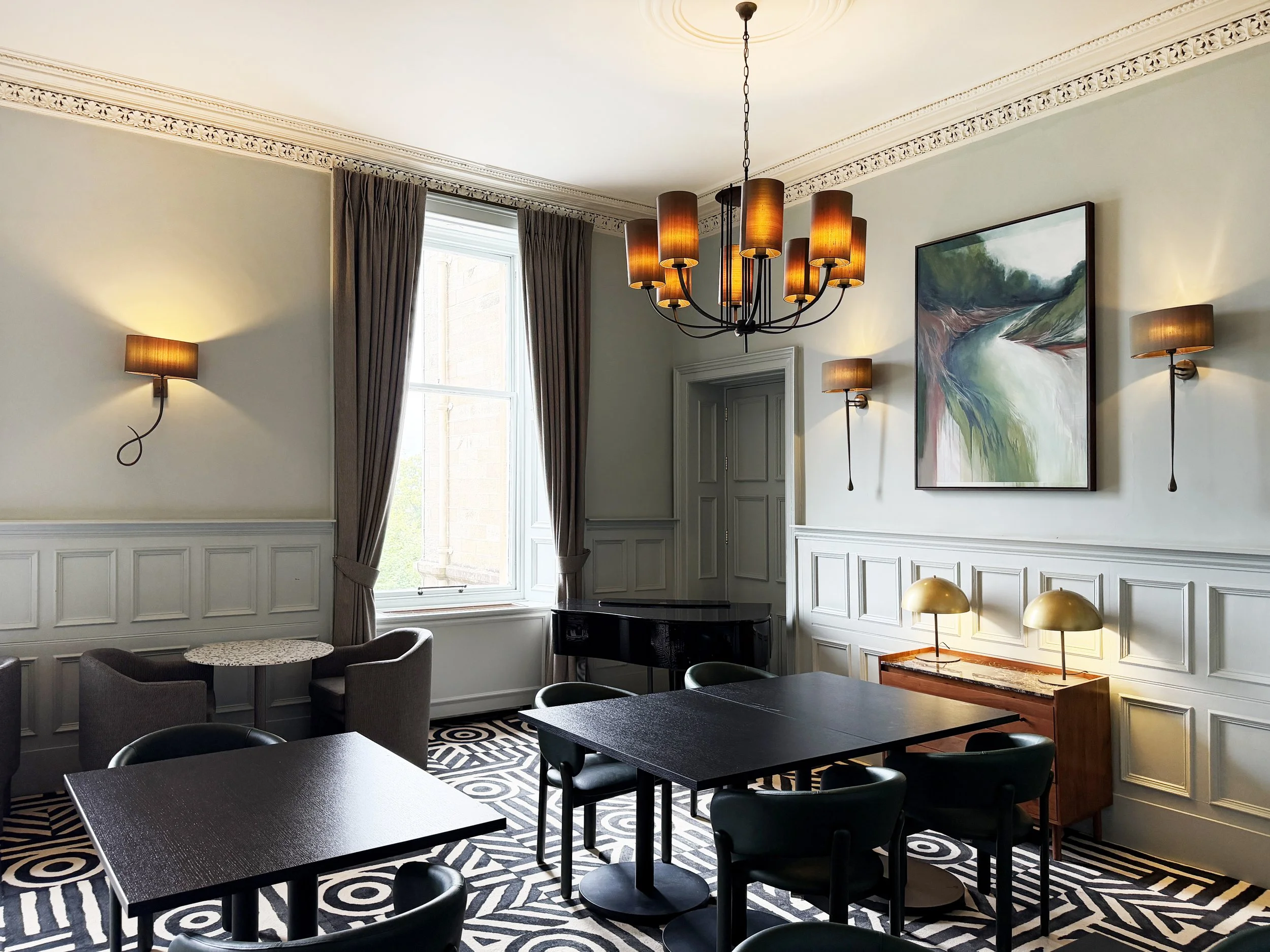 Dunblane Hydro  with elegant molding and wainscoting, a large window with curtains, a modern chandelier with multiple warm-colored cylindrical lamps, wall-mounted lamps, a large abstract painting, black tables, and chairs, with patterned carpet.