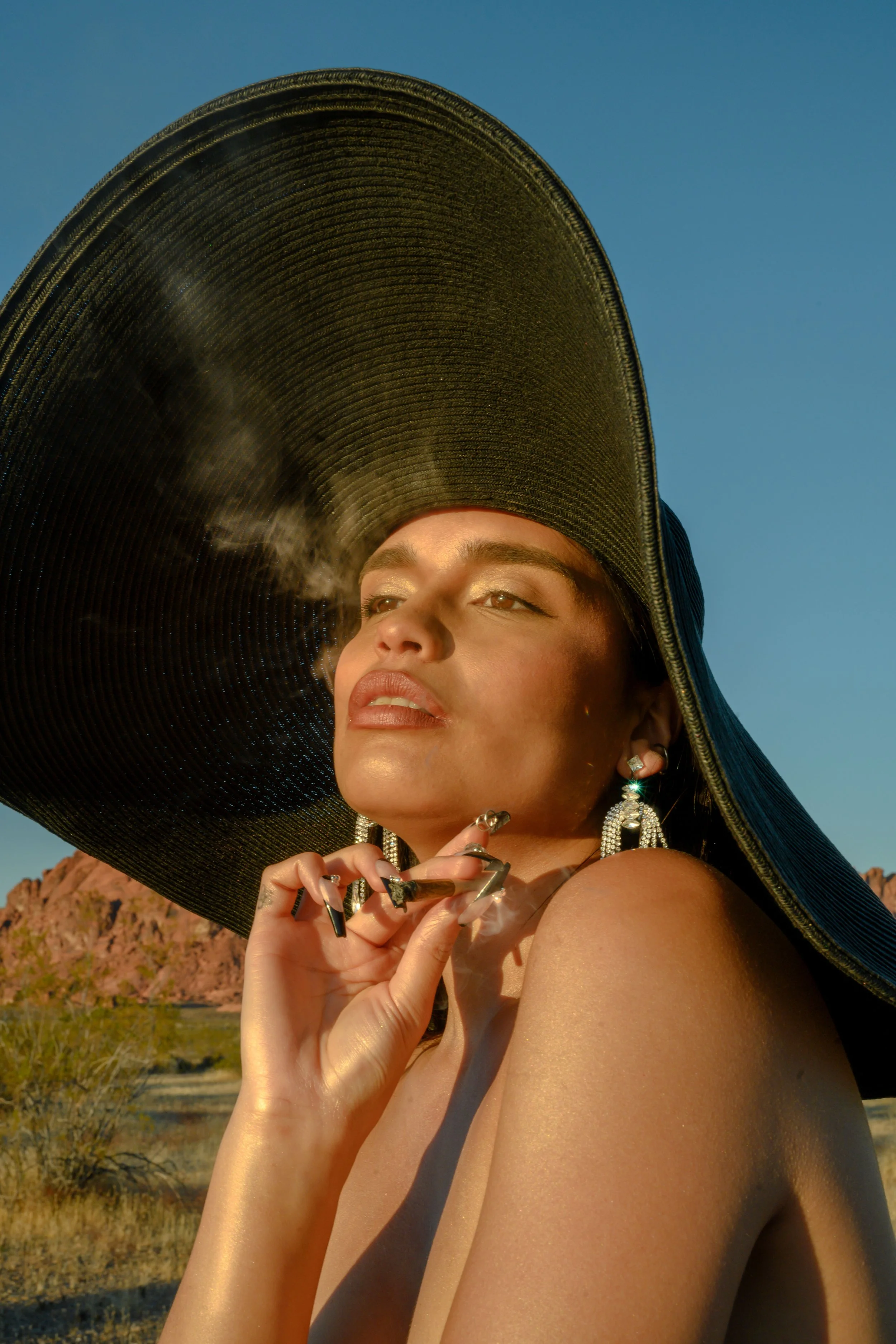 A woman with a large black hat featuring a wide brim, standing outdoors in a desert landscape with rocky hills and sparse vegetation, her face partially visible with jewelry and makeup, with the sky clear and blue in the background.