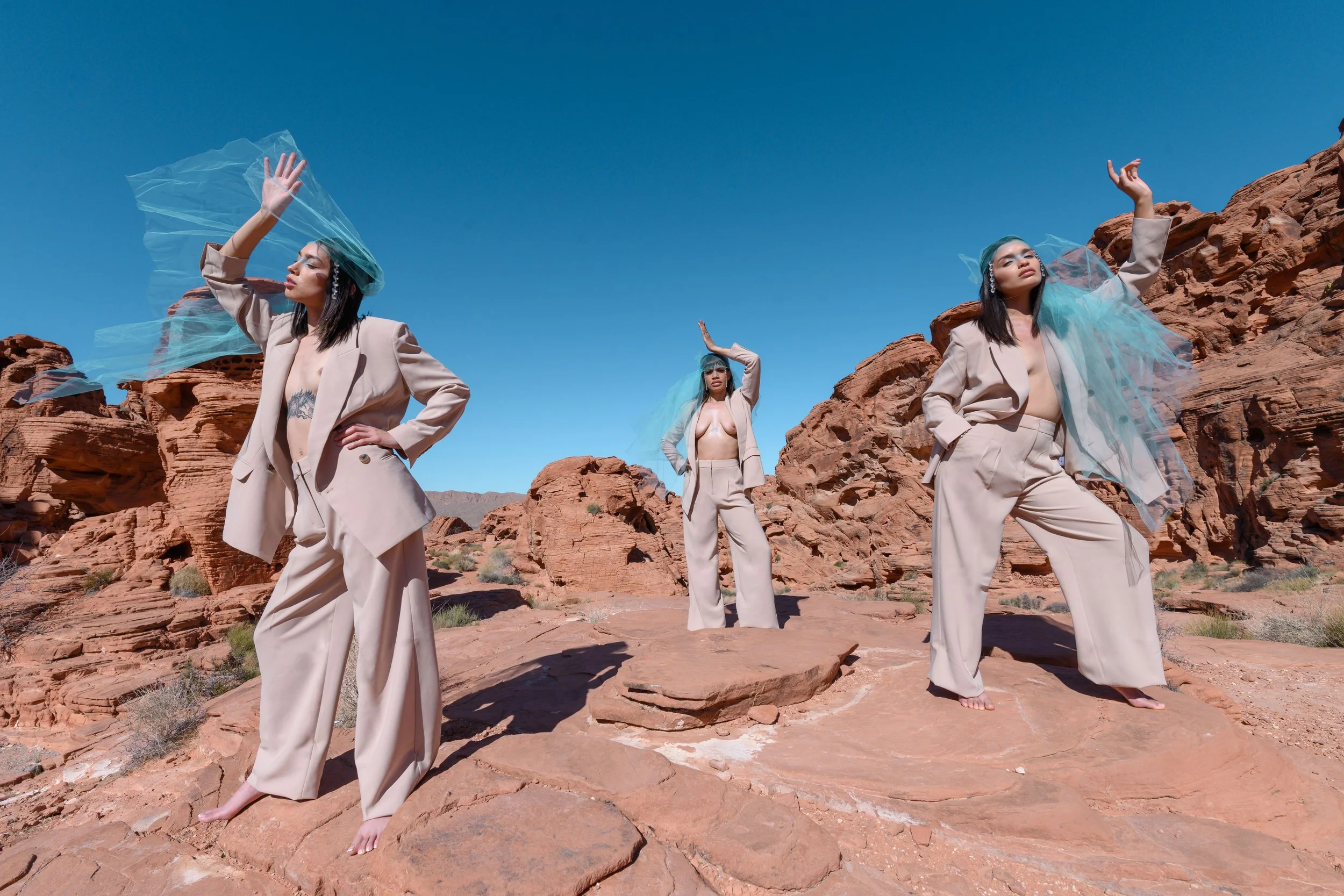 Three women with long black hair standing among red rock formations against a clear blue sky, wearing beige suits with tulle accessories and posing artistically.