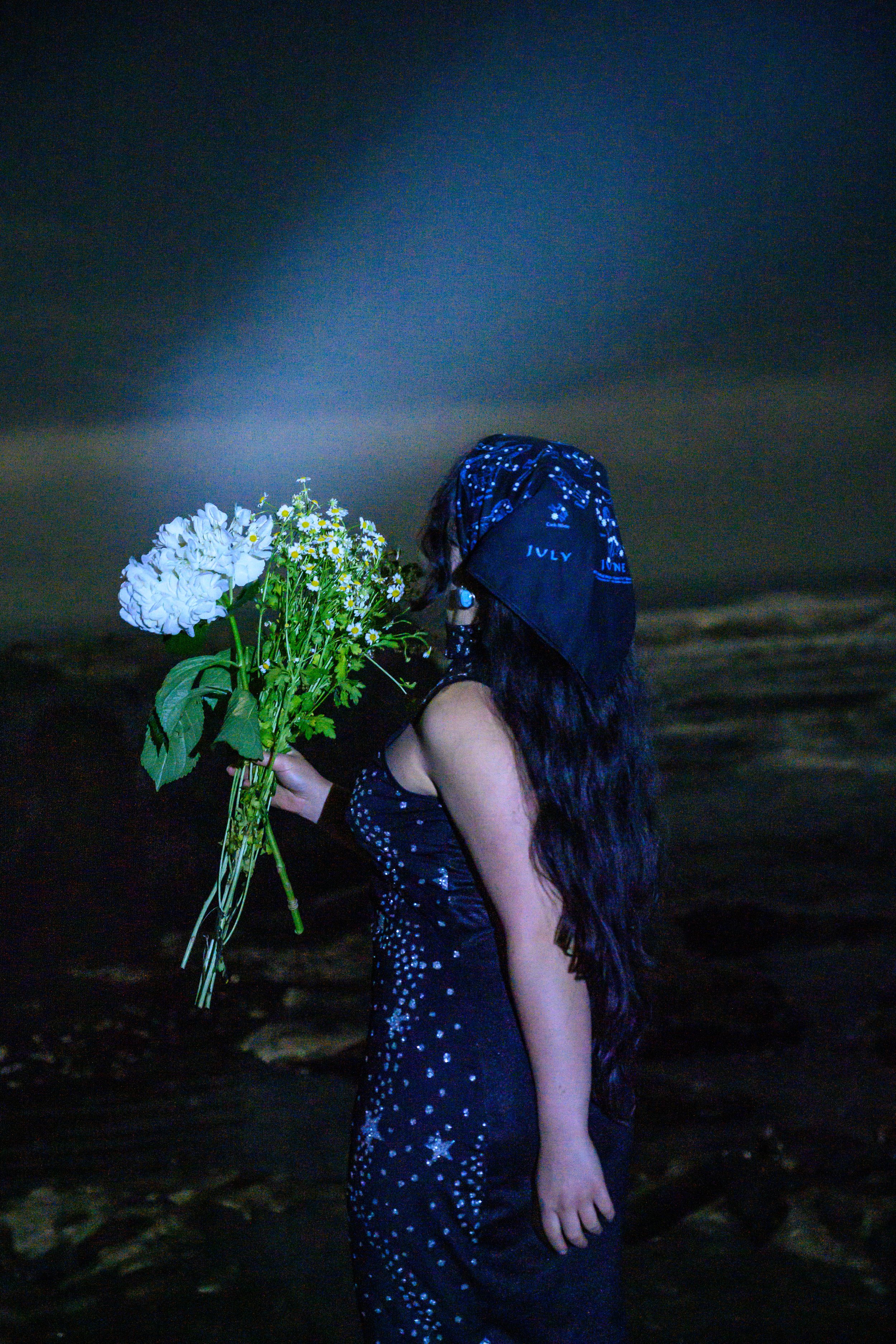 A woman with long dark hair, wearing a black dress with white floral patterns and a blue bandana, holds a bouquet of white flowers, standing in a dark, possibly nighttime setting by the water.