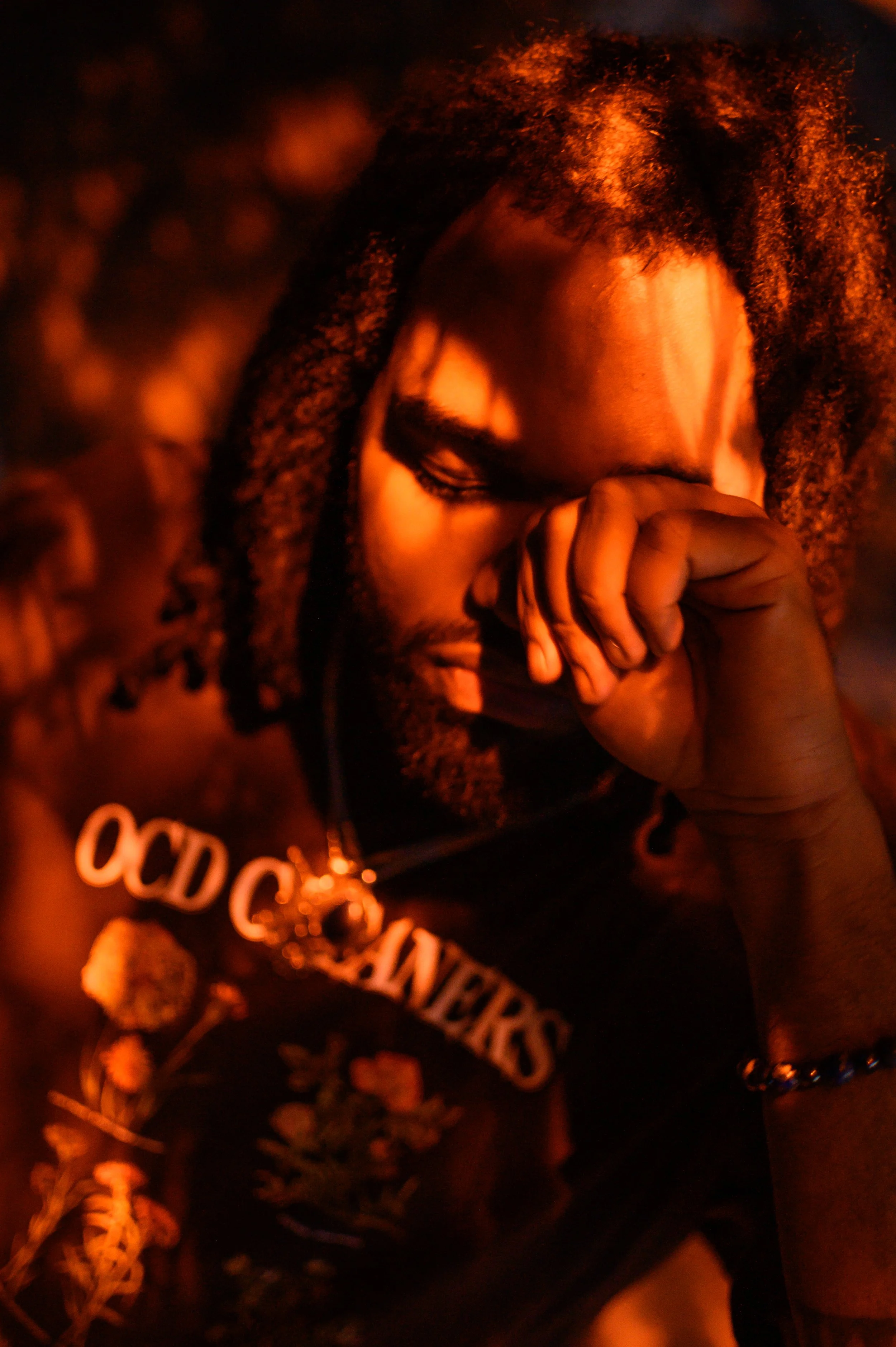 A close-up image of a woman with curly hair, eyes closed, and hand covering her mouth, wearing a black shirt with orange text and jewelry, under warm, reddish lighting.