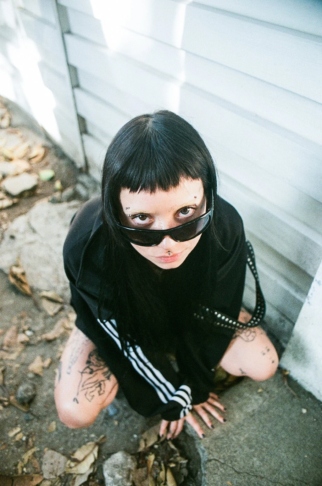 A woman with black hair, tattoos, and piercings, wearing sunglasses and a black outfit with white stripes on the sleeves, crouching on the ground outside near a white siding building with fallen leaves around.