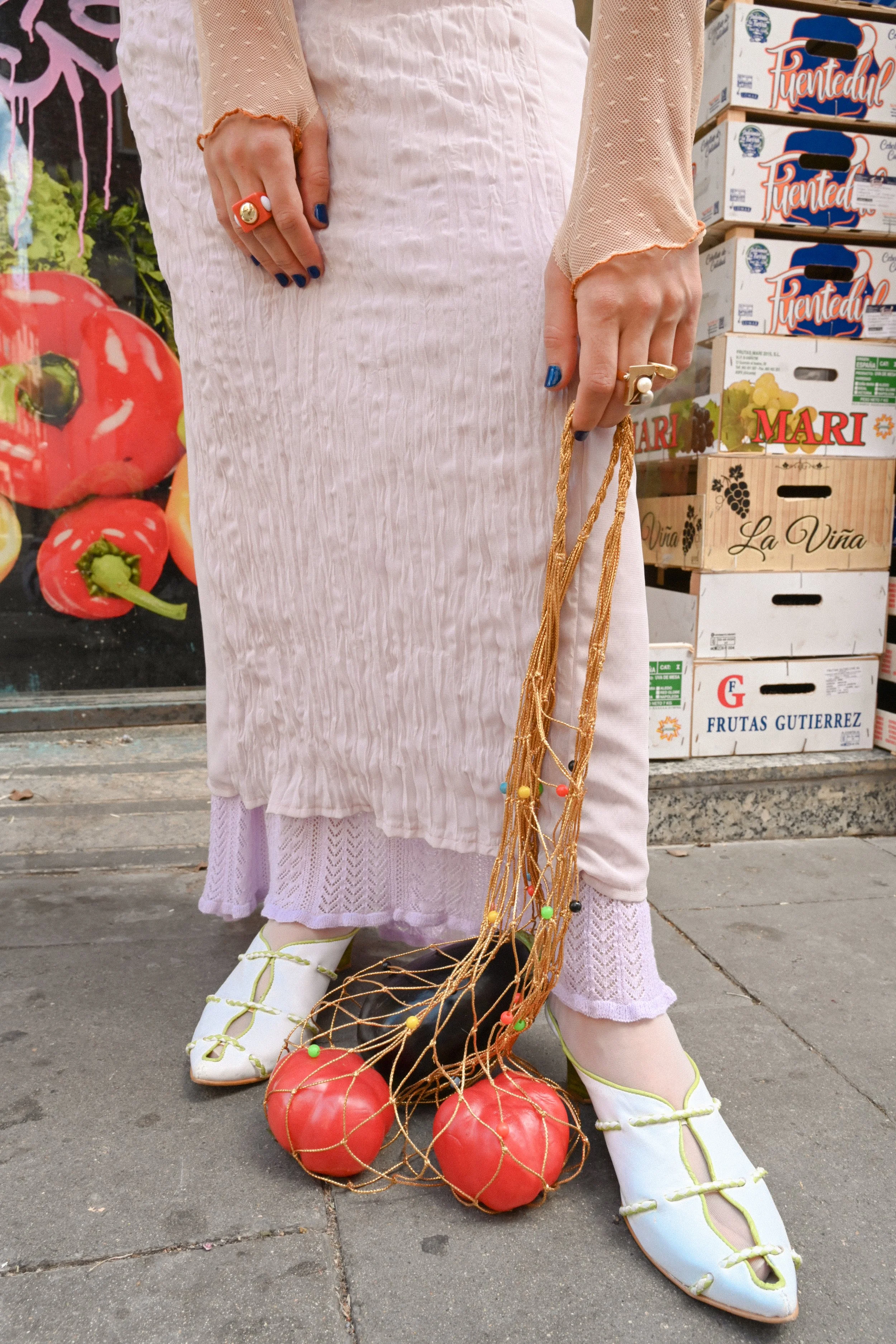 A person dressed in vintage-style clothing standing outside on a sidewalk, holding a rope net filled with large red tomatoes. The person is wearing white lace-up shoes, white pants, a pink and white dress, and a beige long-sleeved top with mesh sleev