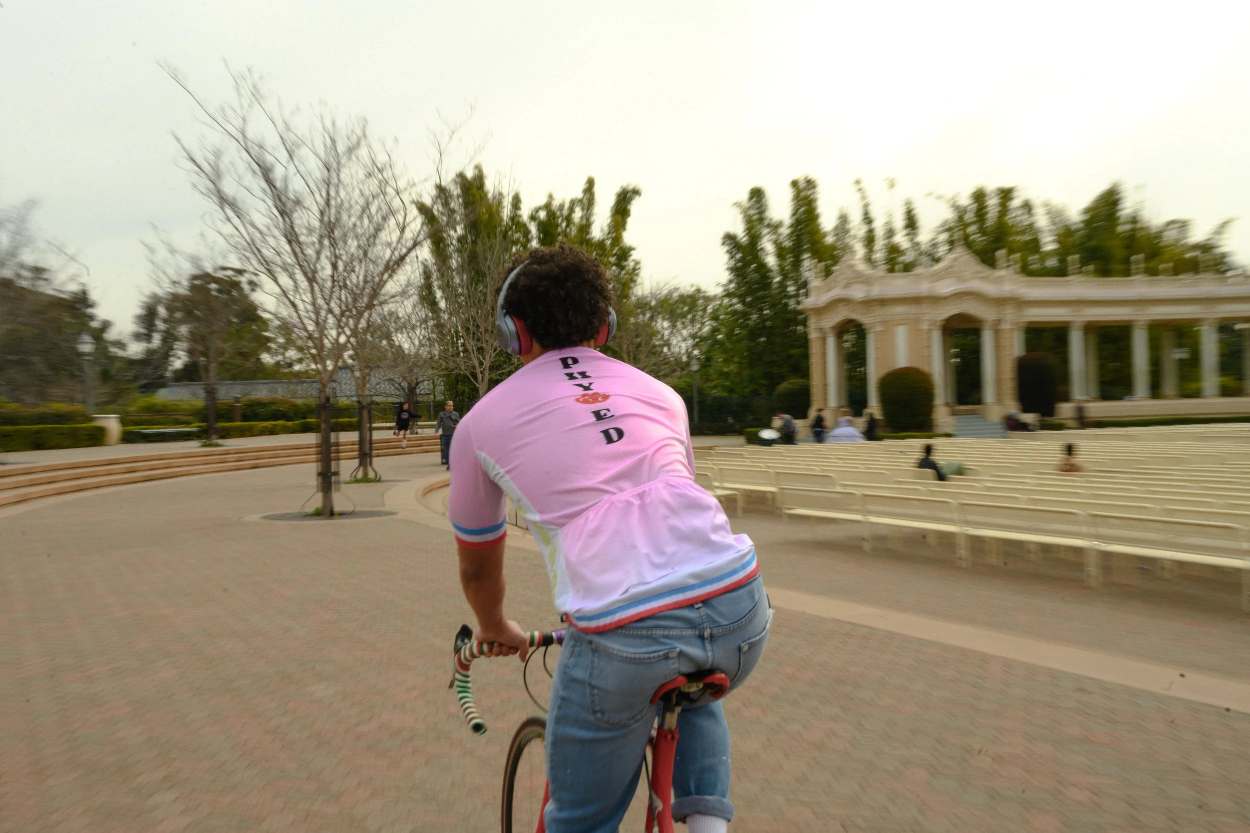 A person riding a scooter in a park, wearing a pink shirt, jeans, and headphones. The park has trees and a pavilion in the background.