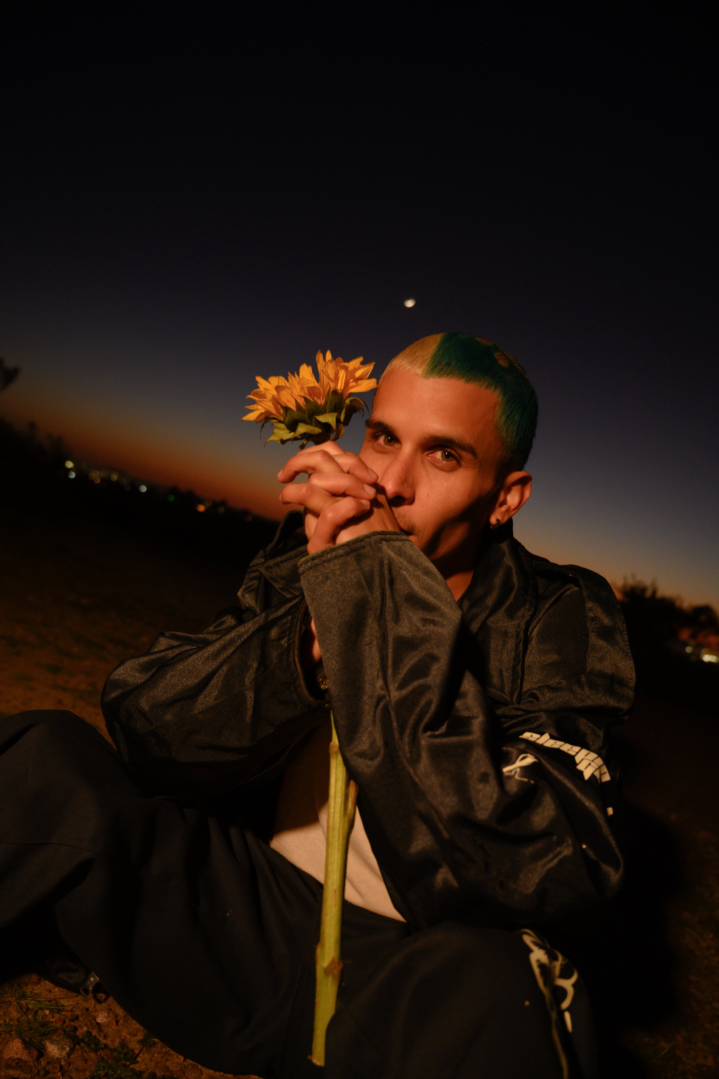 Person with green highlights in hair holding a sunflower in front of their face during dusk or night