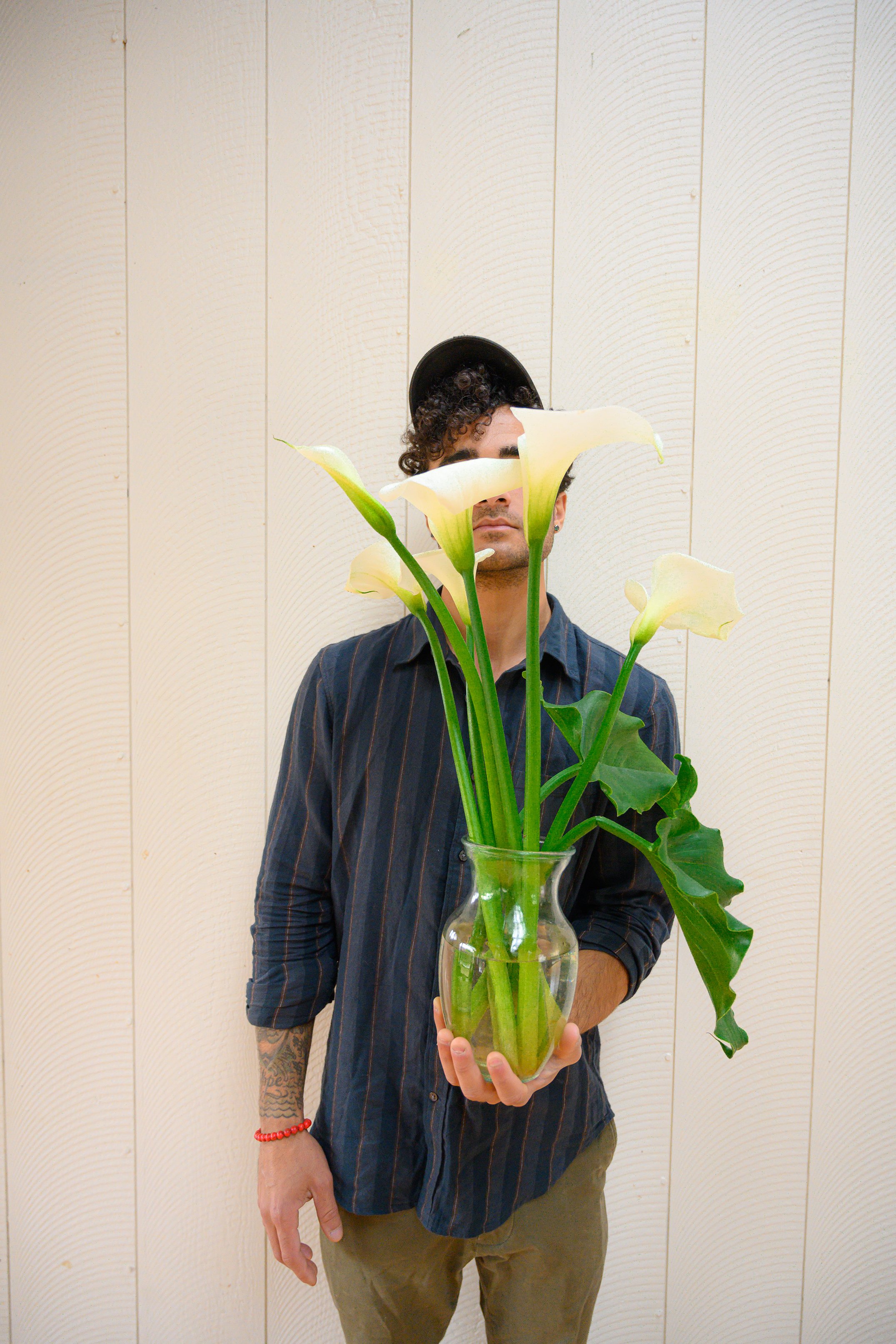 A person with curly hair, wearing a black cap and striped shirt, holding a glass vase with white calla lilies and large green leaves in front of a light-colored wooden wall.