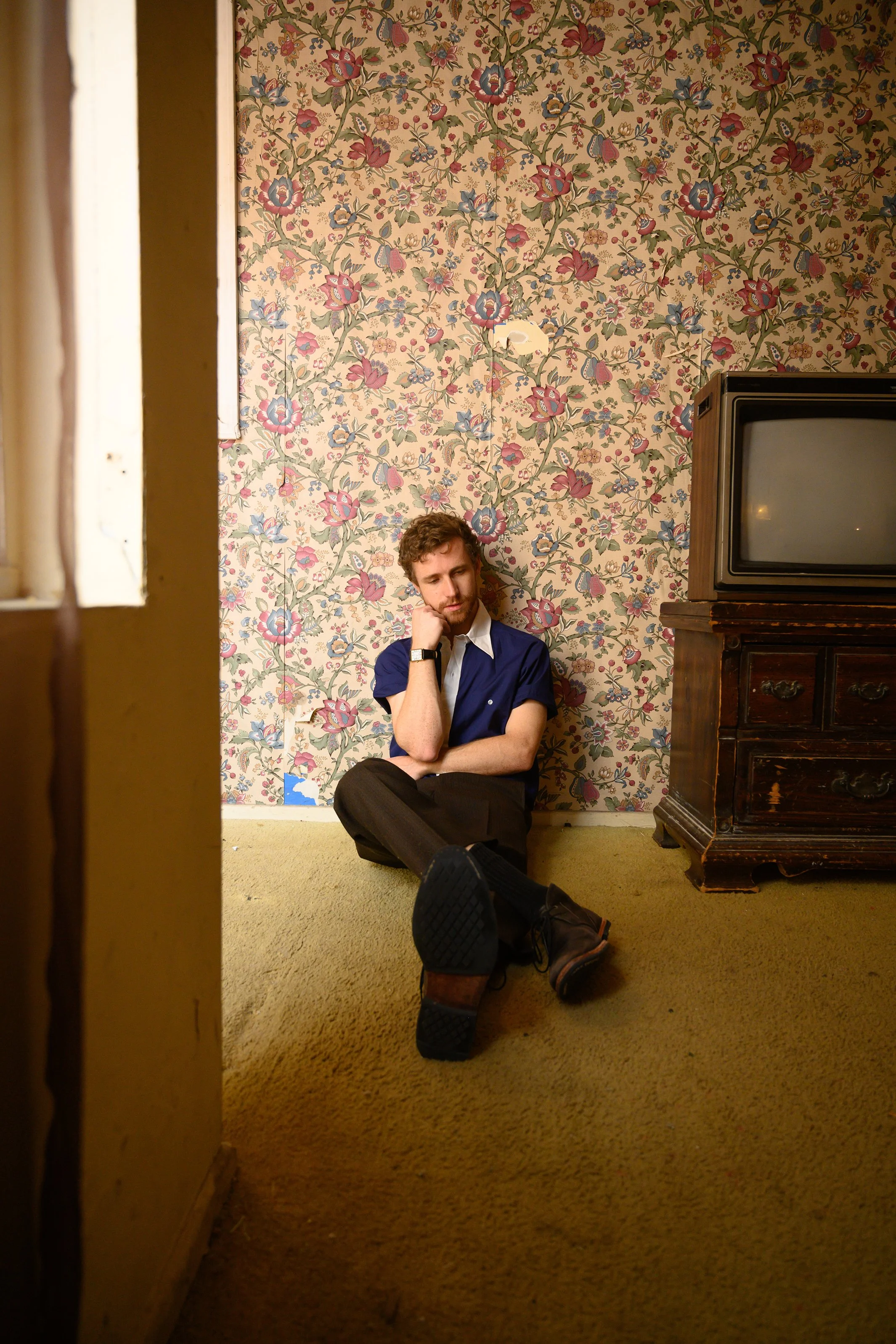A man with curly hair and a beard sitting on the floor in a room with floral wallpaper. He is wearing a navy blue shirt and dark pants, resting his head on his left hand. A vintage television sits on a wooden cabinet to his right. The room has beige 