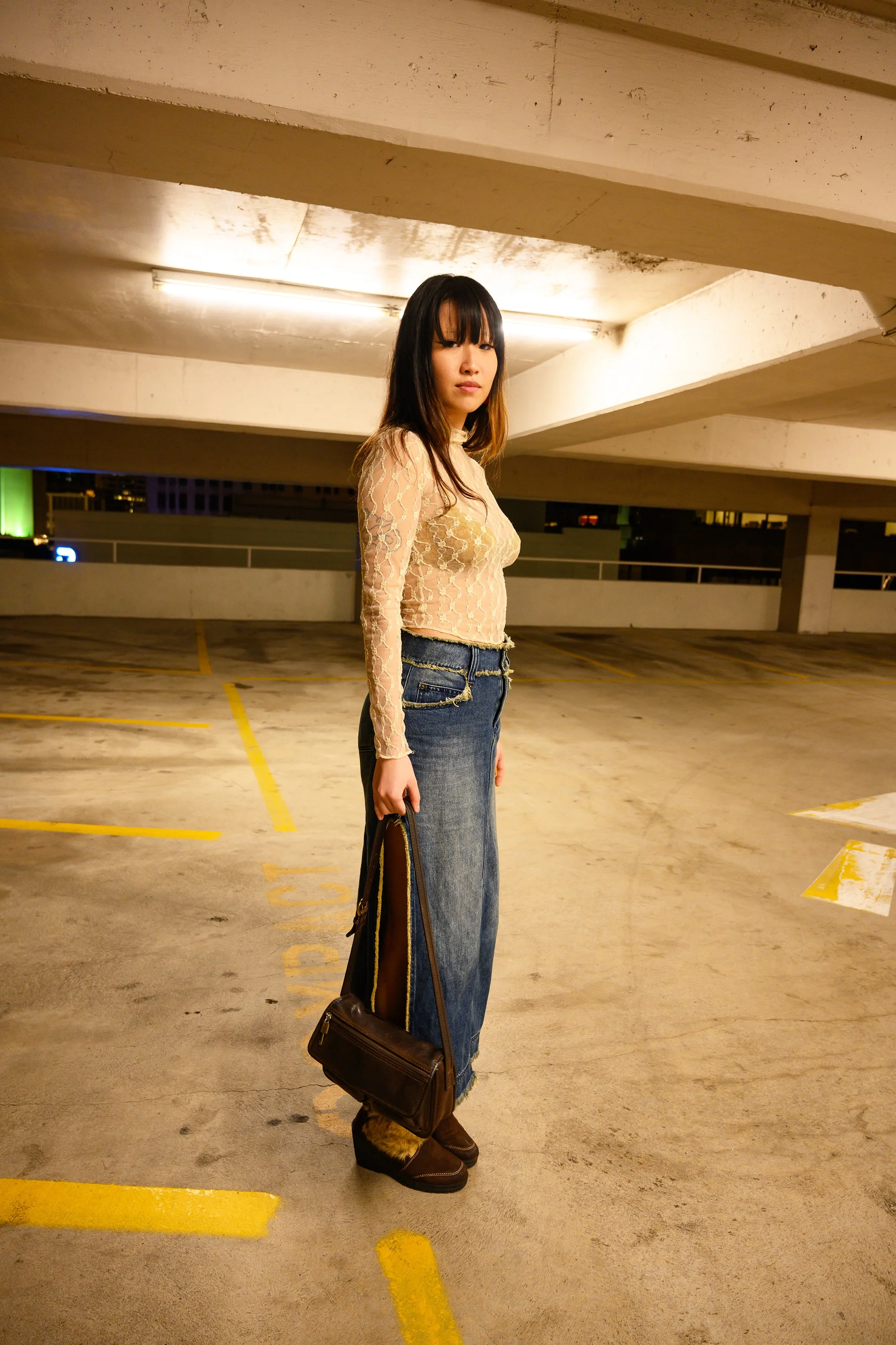 A woman with long dark hair and bangs standing in a parking garage, wearing a beige lace top, wide-leg jeans, brown cowboy boots, and holding a brown shoulder bag.