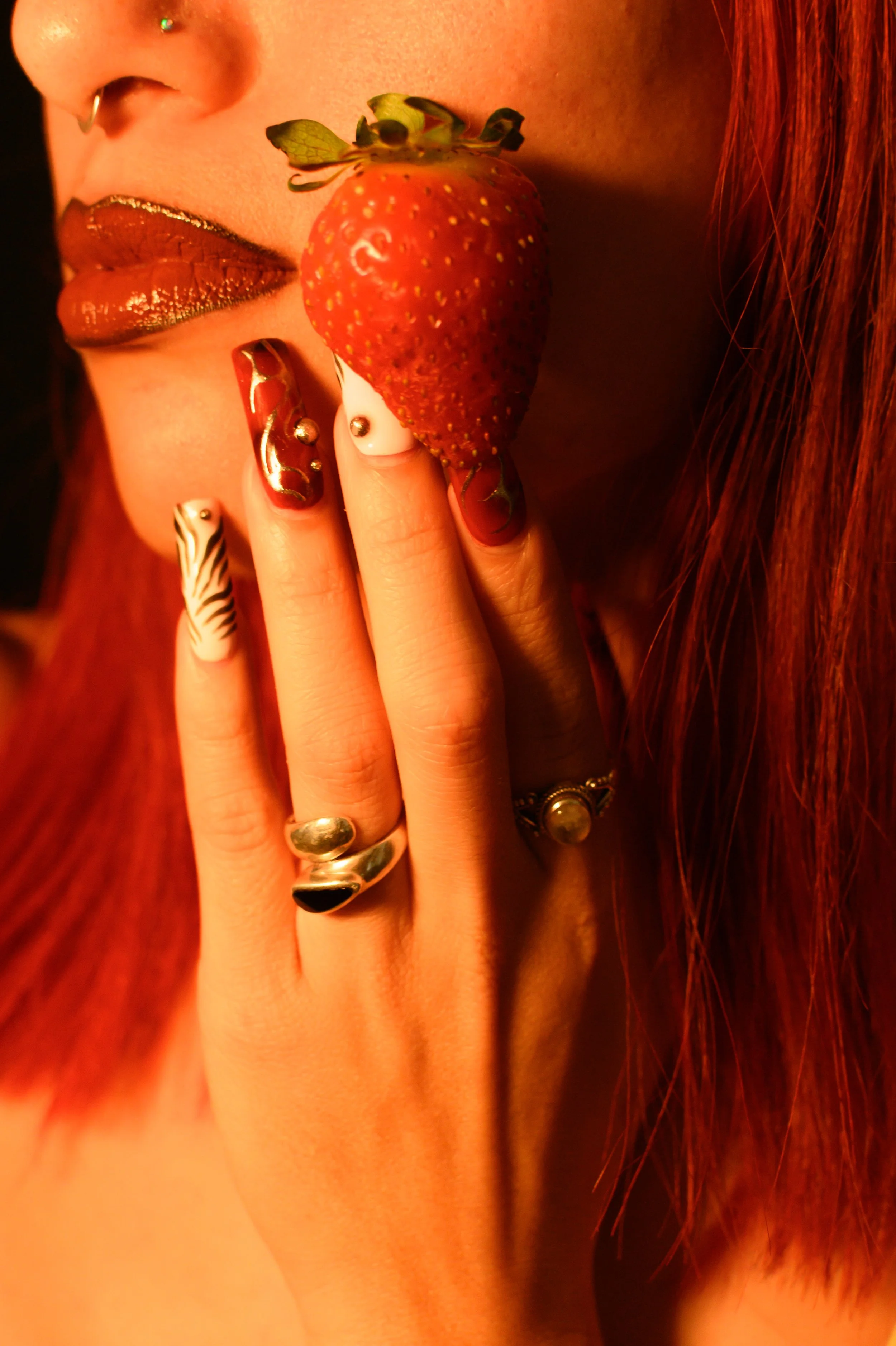 Close-up of a woman with red hair, wearing makeup with metallic lips and nails, holding a strawberry near her face.