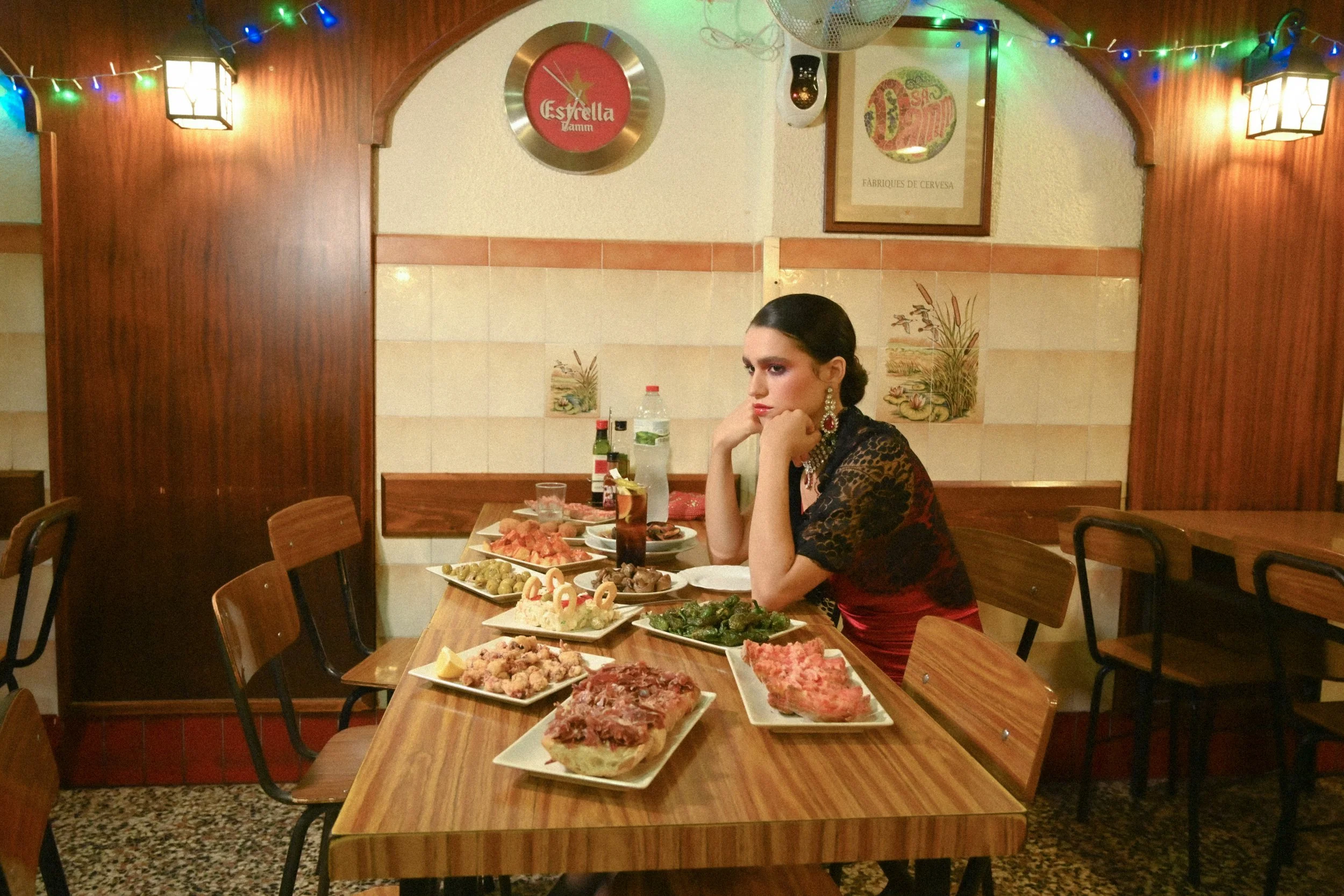 A woman wearing a dark lace dress and large earrings sits at a restaurant table in front of an assortment of dishes, including various meats, vegetables, and appetizers, with bottles and glasses on the table. The restaurant decor features wooden wall