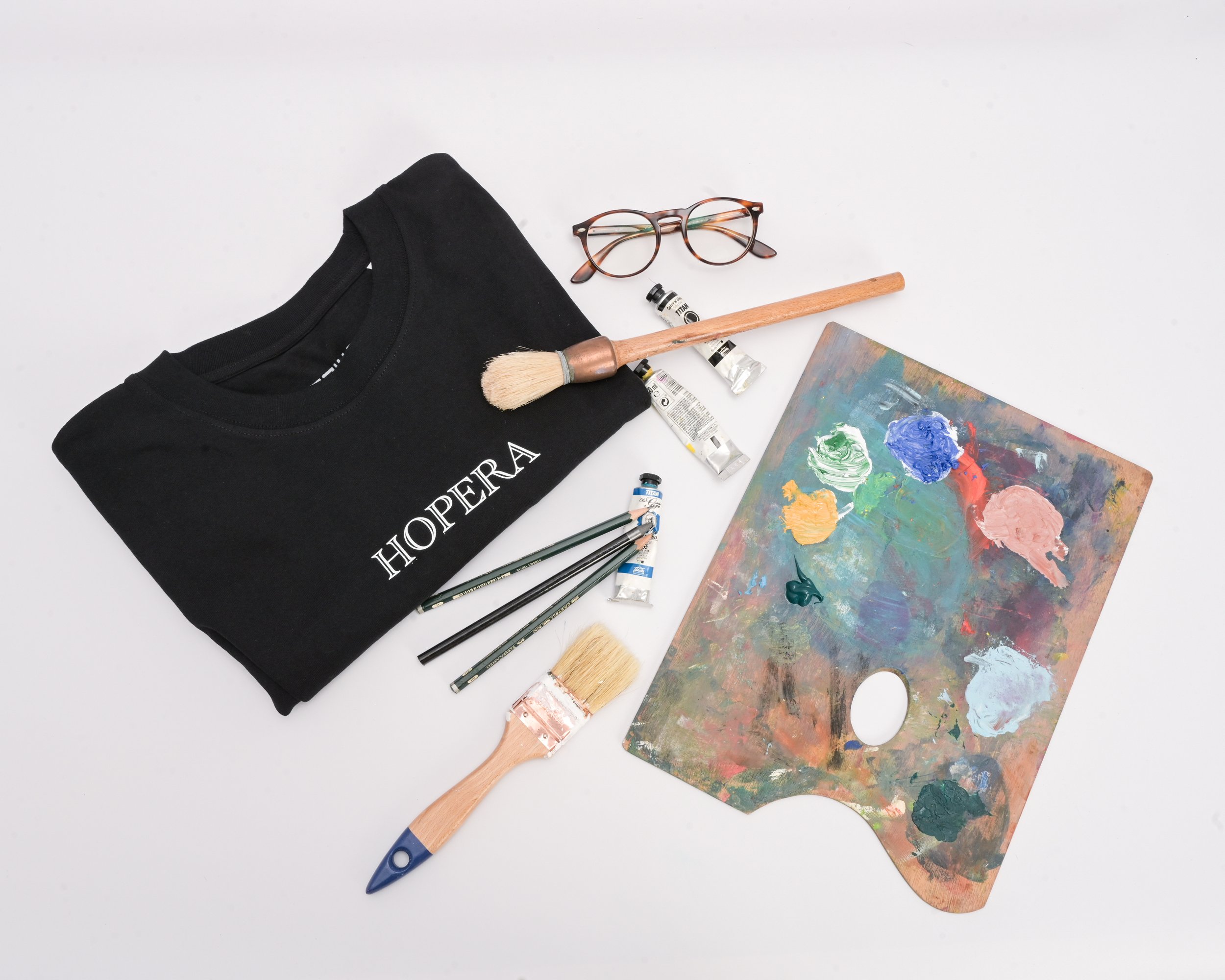 Flat lay of art supplies including a black t-shirt with white text, paint tubes, a paintbrush, a palette with paint, glasses, and paint pens on a white background.