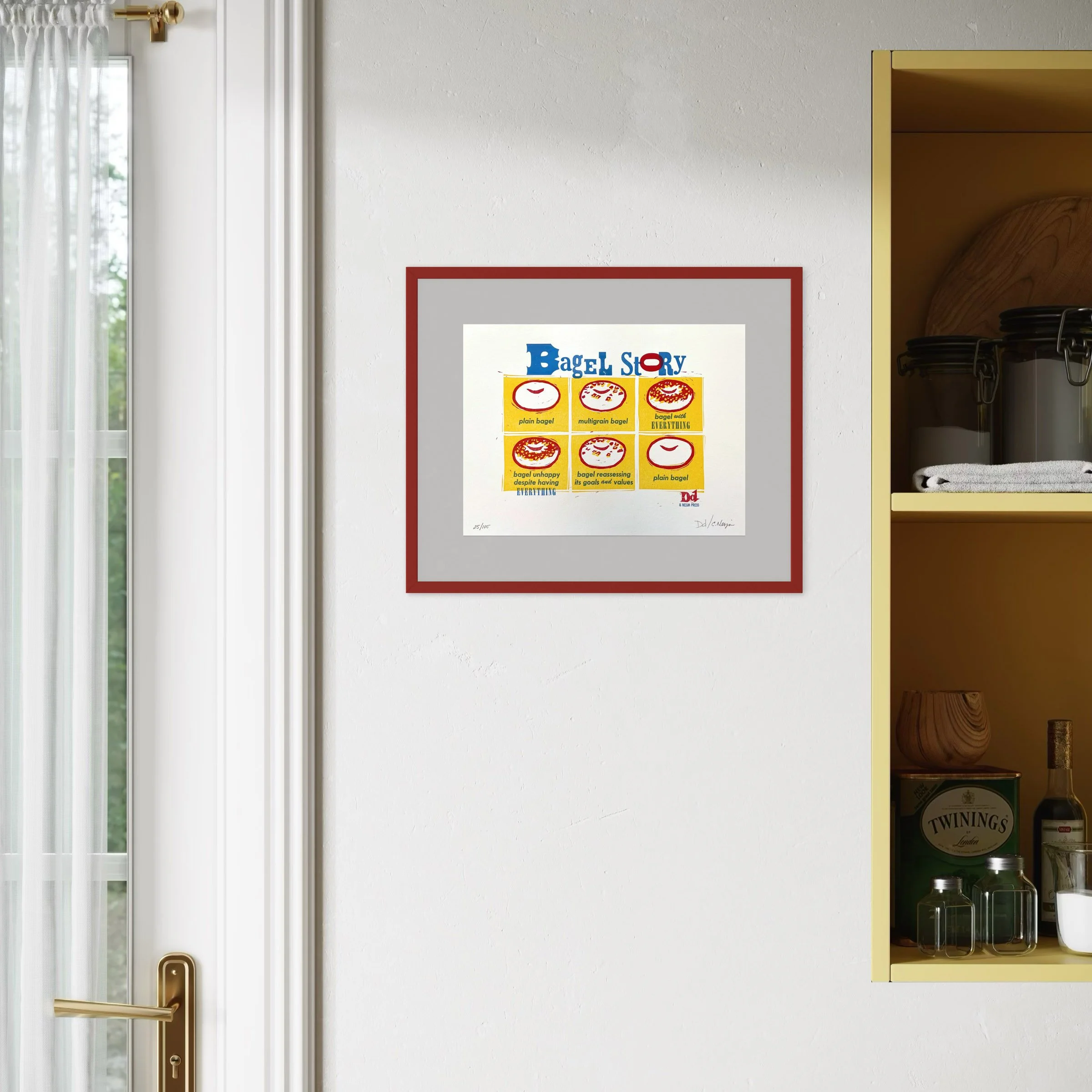 Framed example of 'Bagel Story' letterpress and linocut art print hanging in a kitchen or dining area setting. The print is shown framed with a light gray mat and a dark red frame.