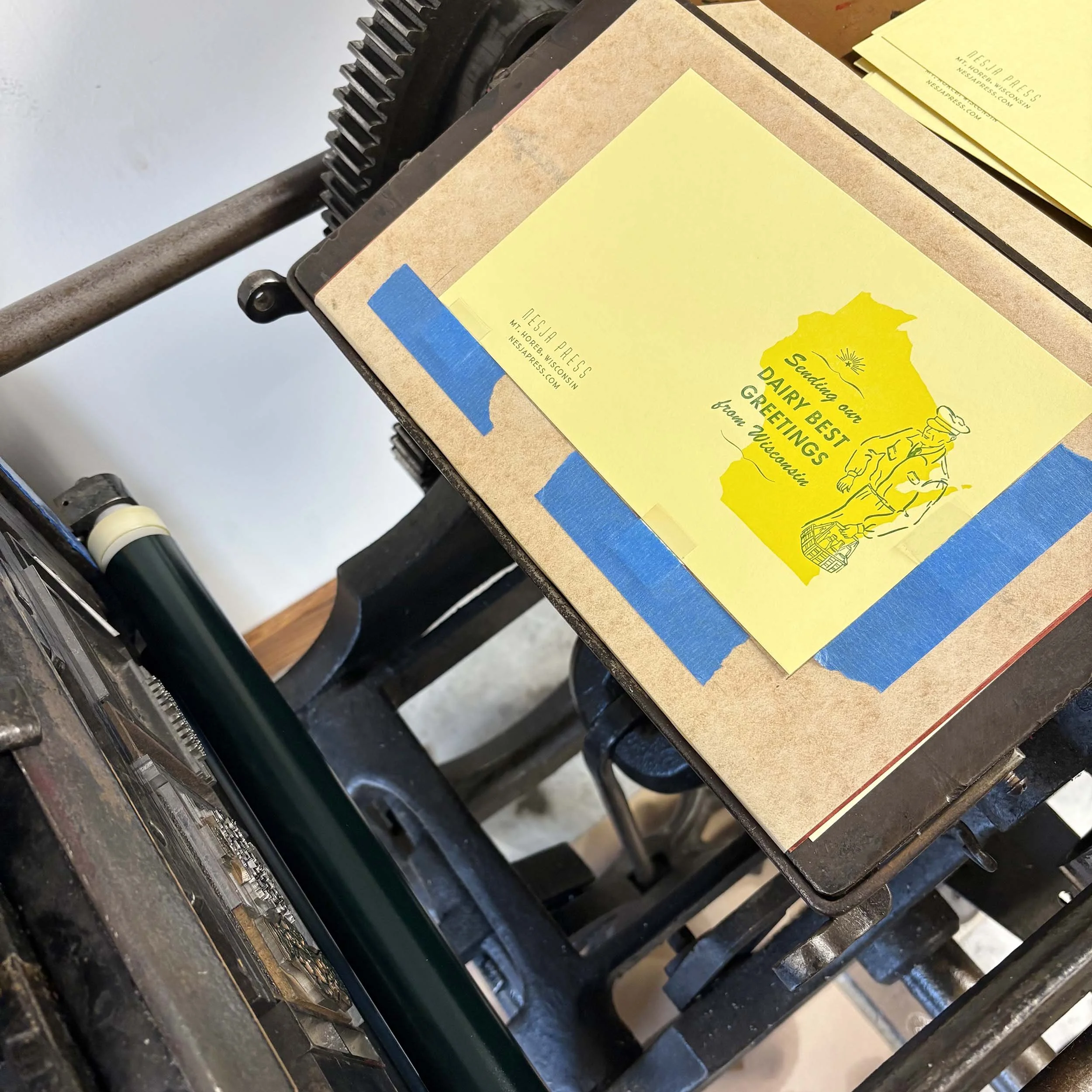 A 'dairy best greetings' card is shown on a vintage printing press. It features the shape of Wisconsin in yellow on yellow paper. Freshly printed green type and a milk man graphic overlay the state.