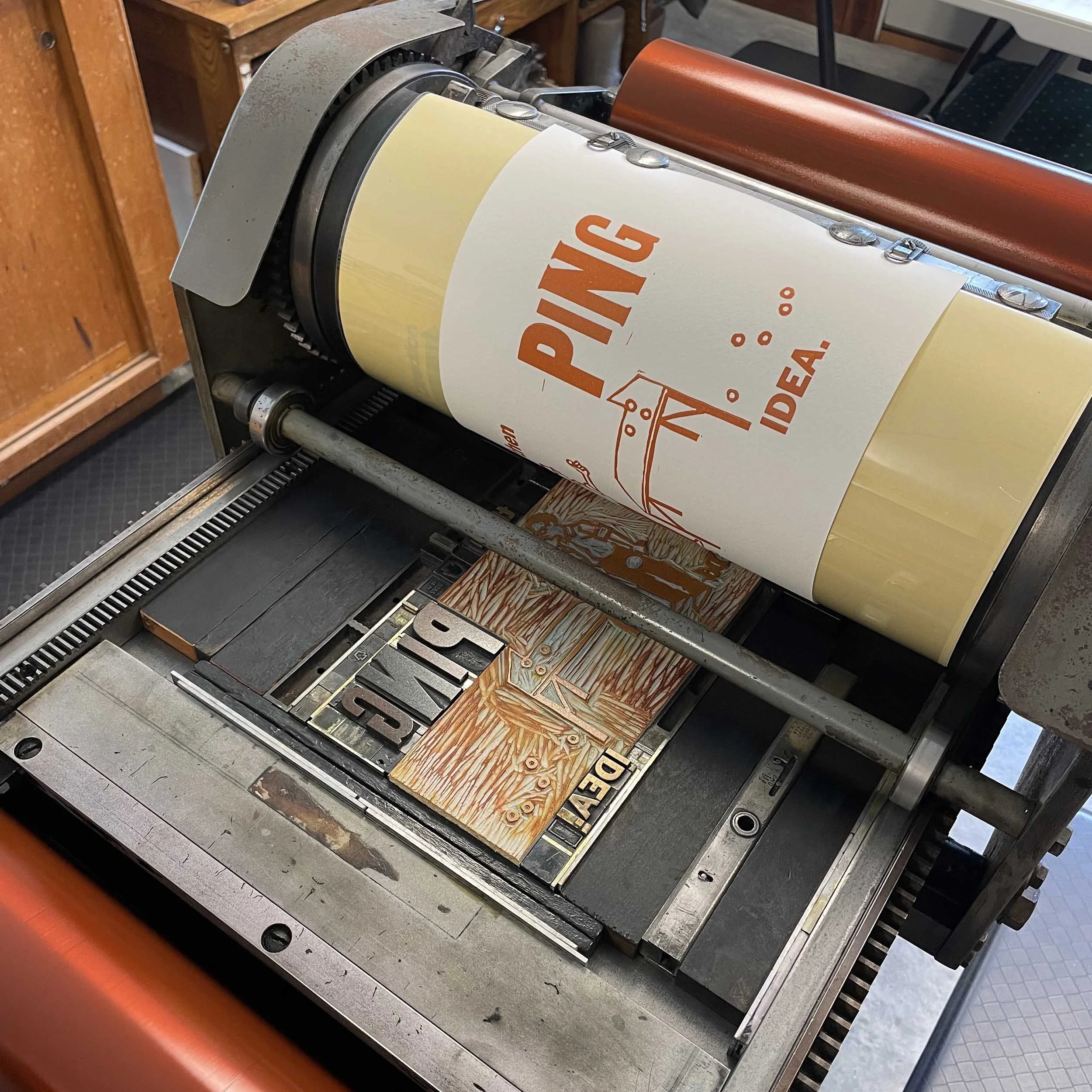 Close-up view of a vintage printing press printing orange ink on white paper. The image shows the frame of a ping pong table and text with the words 'Ping' and 'Idea.'