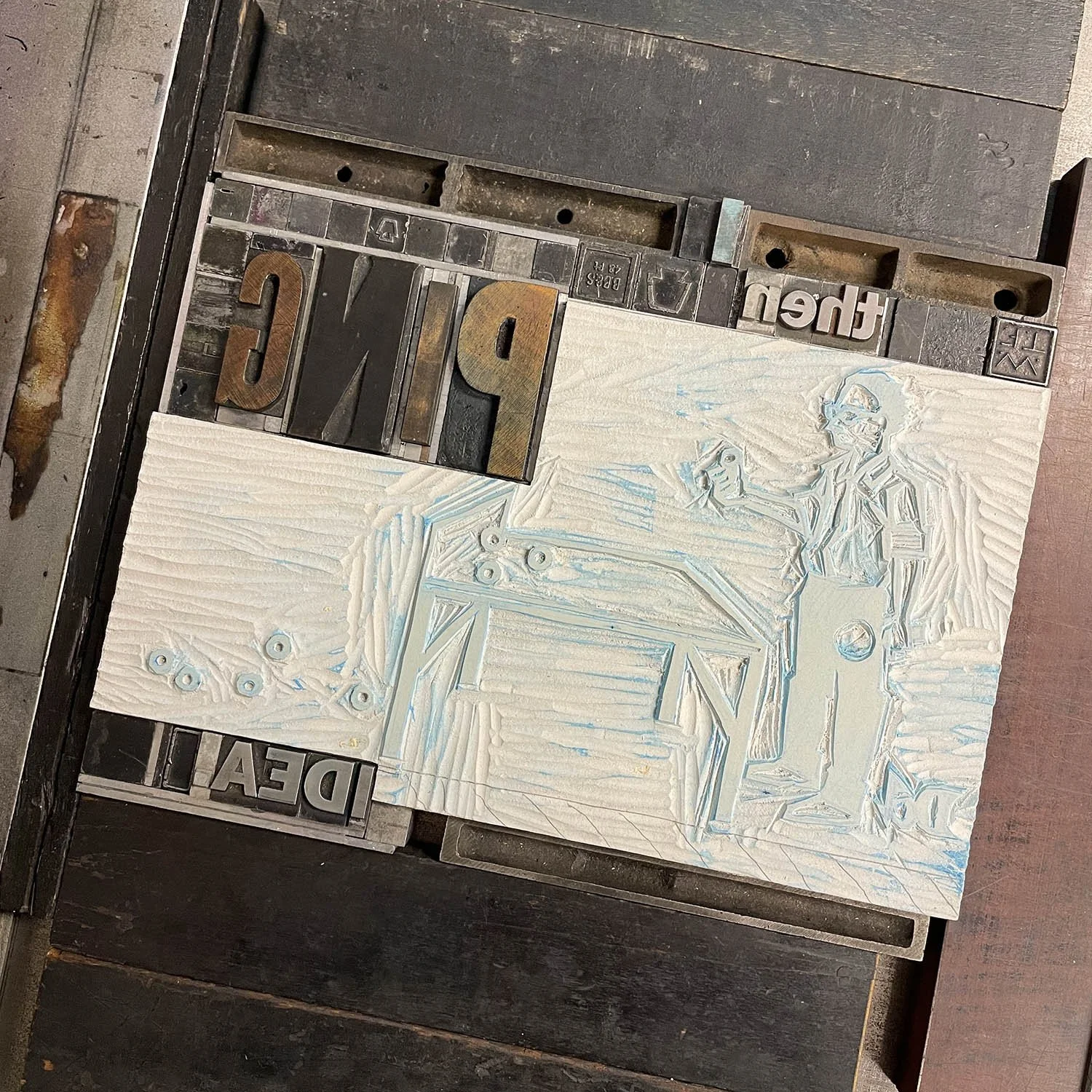 Overhead view of a printing press with a linoleum block, wood and metal type. The linocut image depicts a person at a table with small objects. The metal type includes the words 'PING' and 'IDEA'.