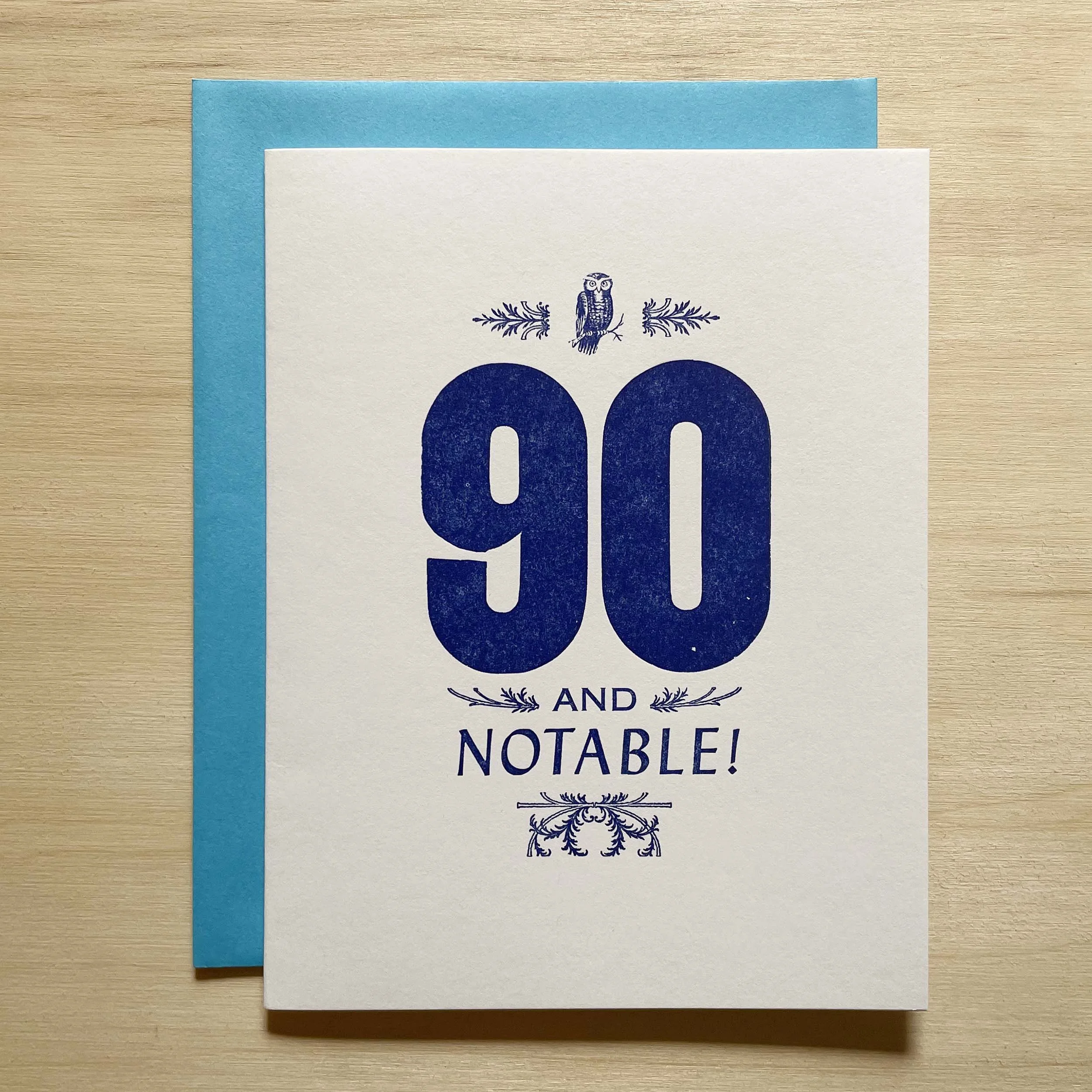 90 and Notable card features a bold 90 with smaller words AND NOTABLE. Letterpress ornaments add decoratio. Dark royal blue ink on white paper. Bright blue envelope.