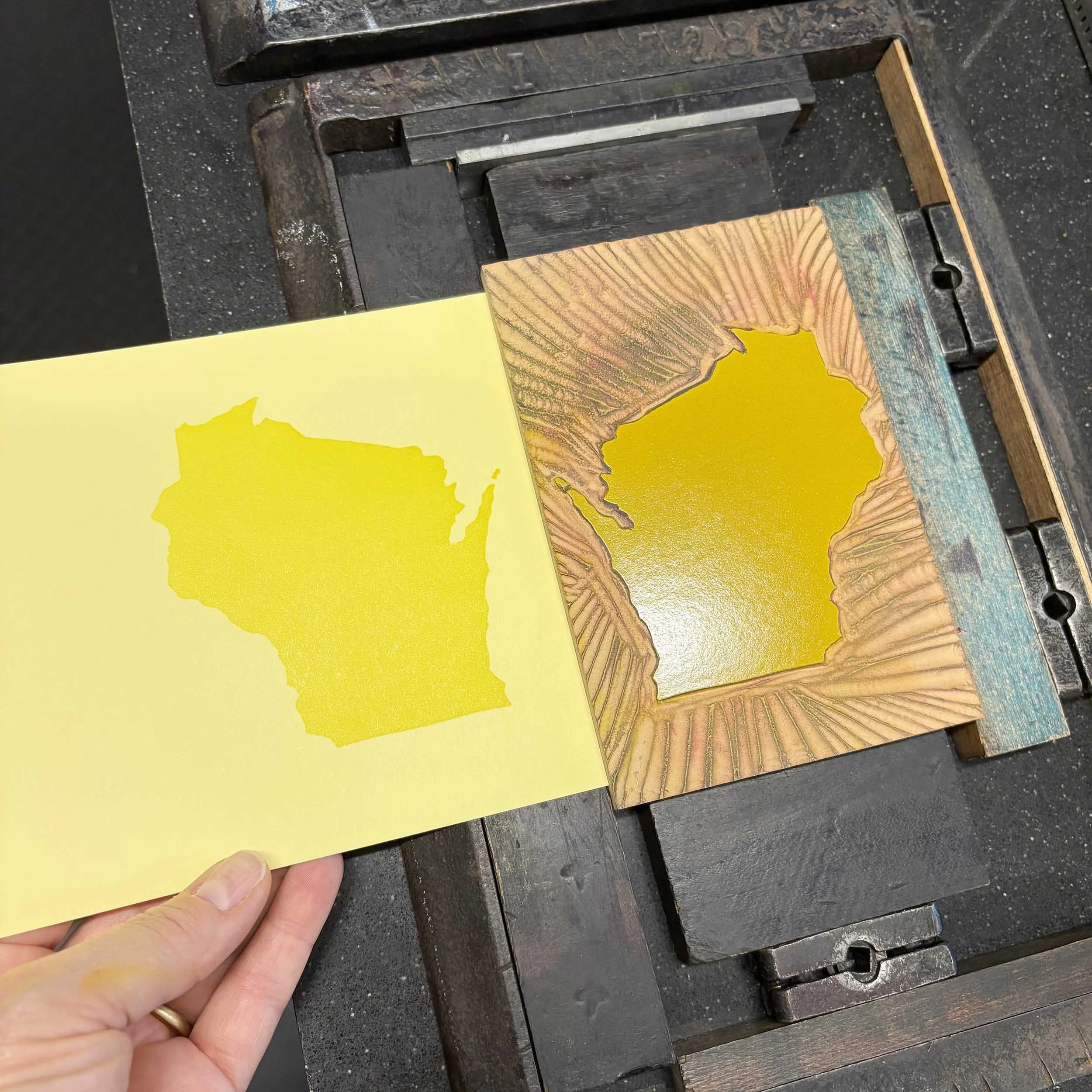 A linocut of Wisconsin is locked in the bed of a printing press. The block is inked. A printed example is held next to it. The Wisconsin shape is printed in dark yellow on light yellow paper.