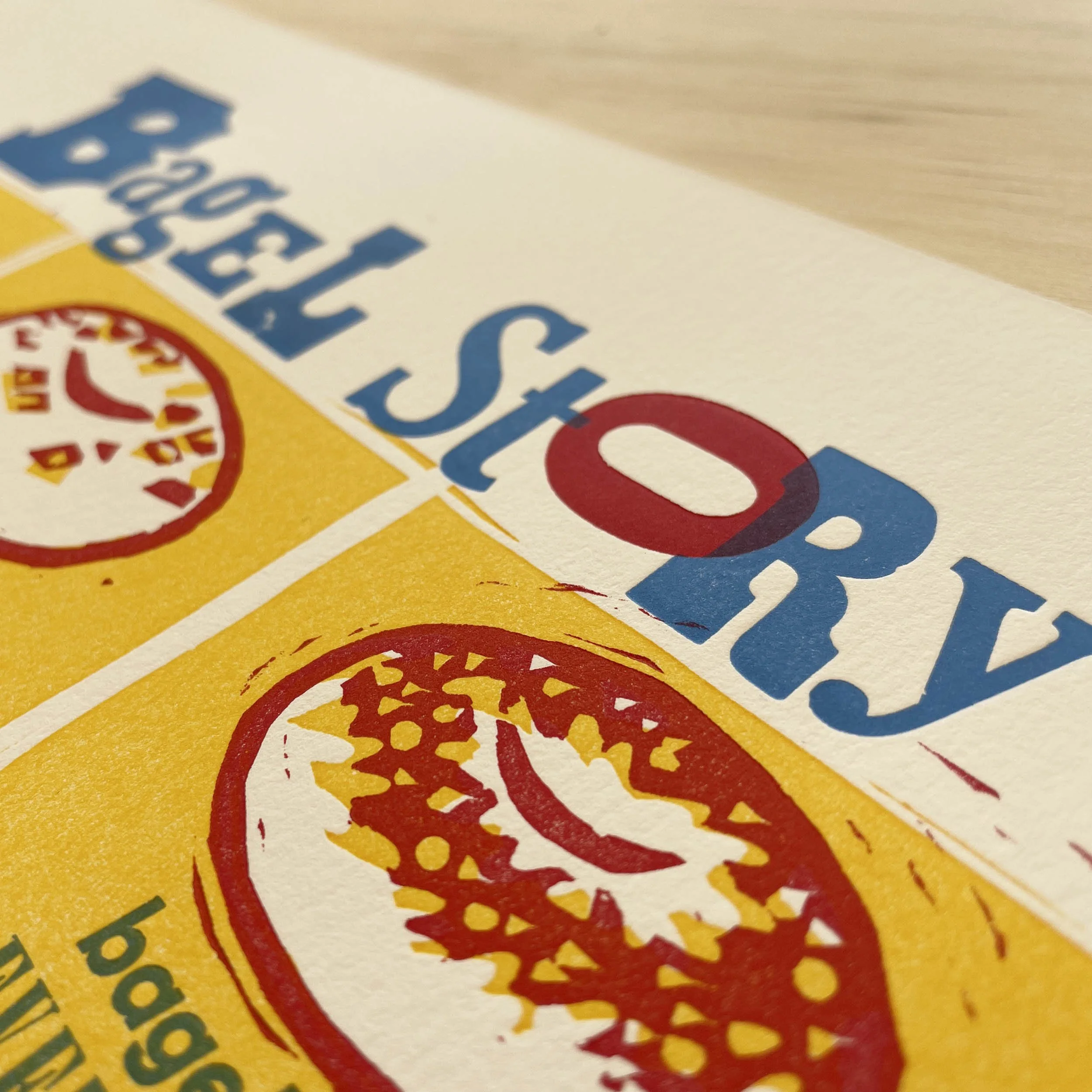 Detail of a six-panel linocut comic follows a bagel’s life. Woodcut-style illustrations in red and yellow panels with handset letterpress type in vibrant blue. Detail features the Bagel Story title.